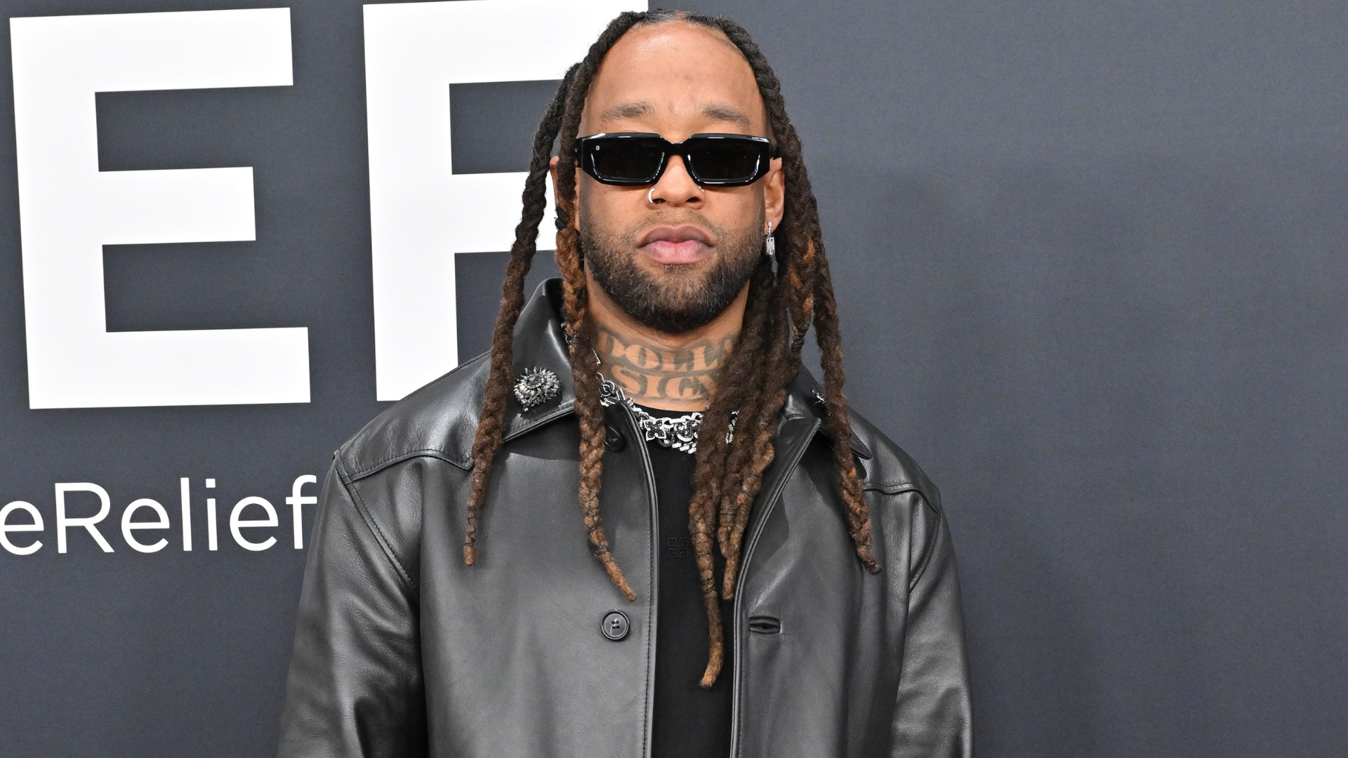 A man with long dreadlocks, sunglasses, and a leather jacket stands in front of a dark backdrop.