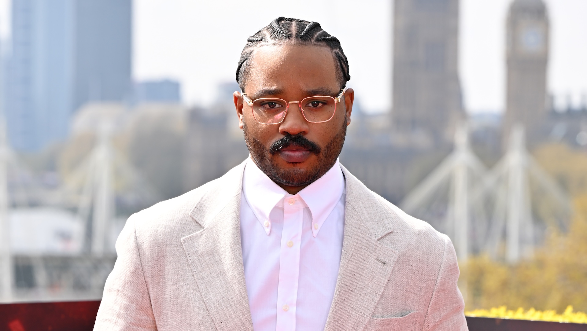 Film director Ryan Coogler is looking at the camera. He is wearing glasses, a white button-down shirt, and an off-white suit jacket. 