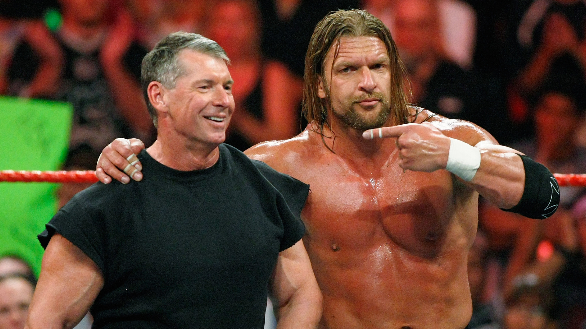 Vince McMahon and Triple H in a wrestling ring, smiling and posing. McMahon wears a black shirt; Triple H is shirtless.