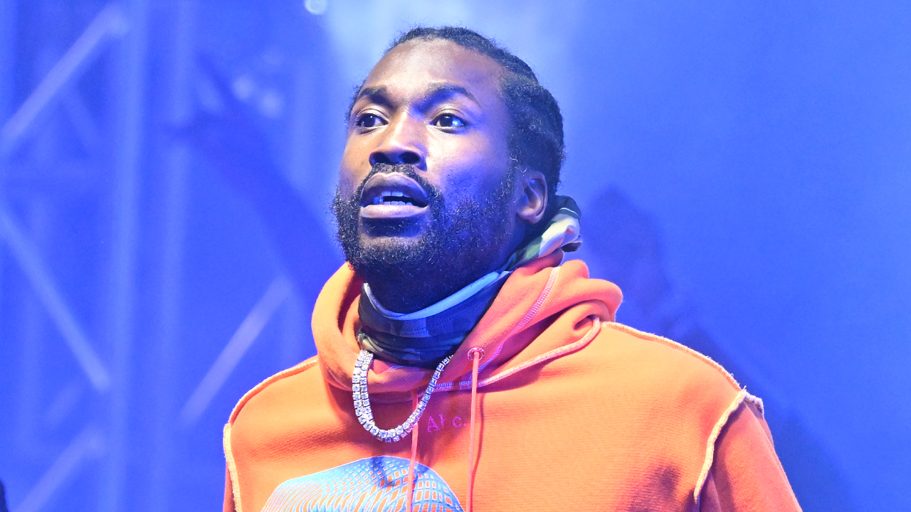 Meek Mill in an orange hoodie with a diamond chain, standing in a blue-lit environment.