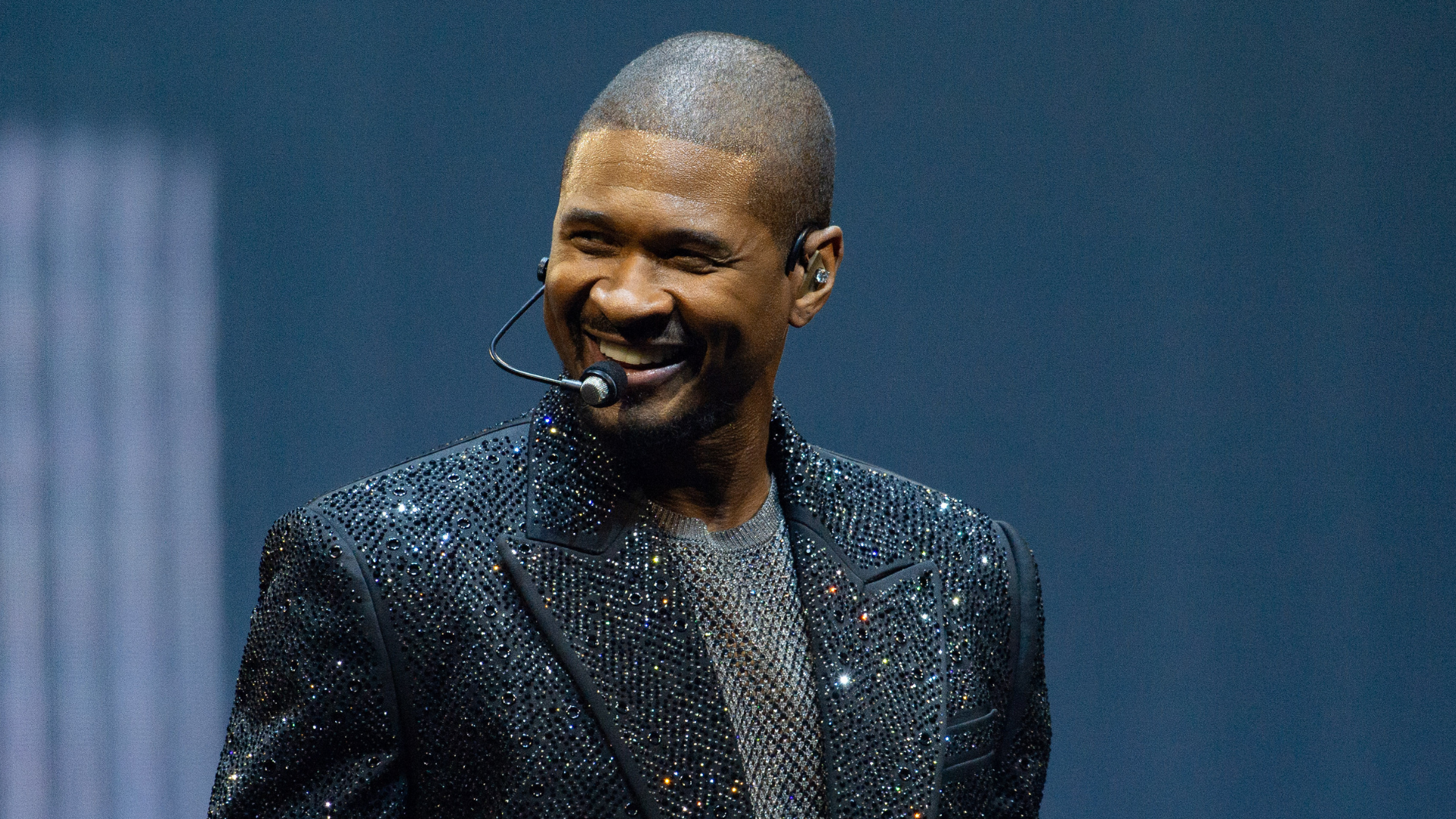 Usher performing on stage, wearing a sparkling black jacket and headset microphone, smiling against a blue background.