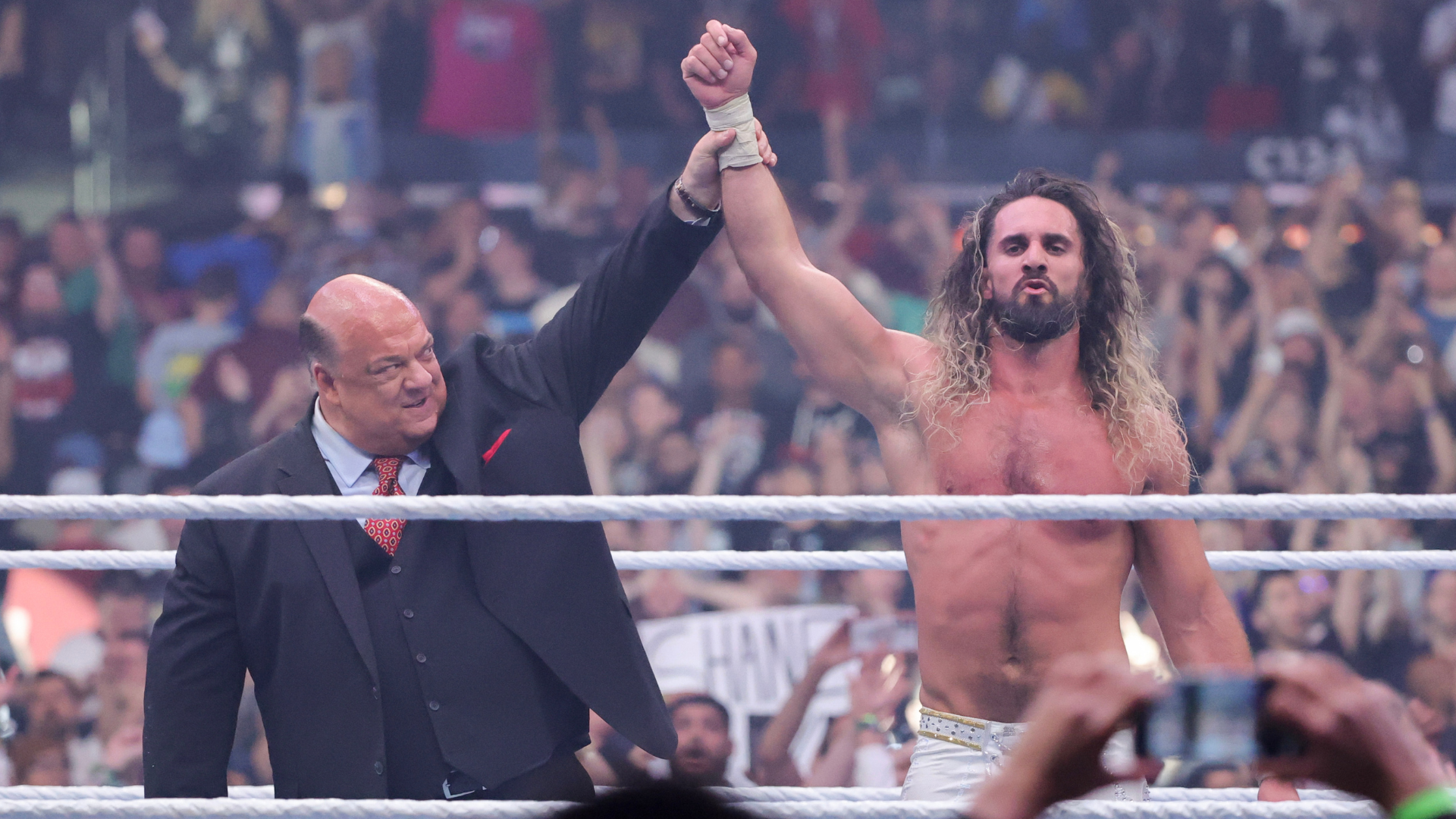 Paul Heyman holds up the arm of a victorious Seth Rollins in a wrestling ring, with a cheering crowd in the background.