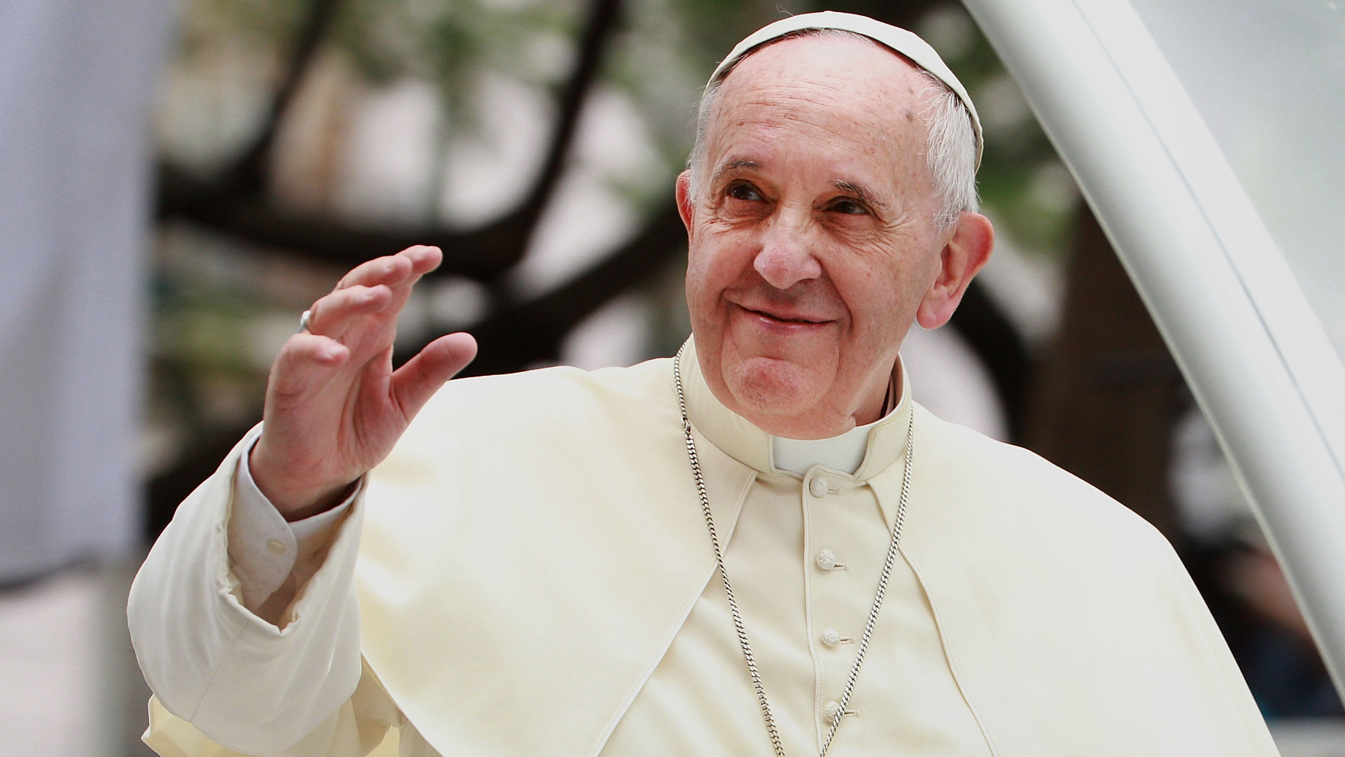 Pope Francis waving, wearing white papal attire and a cross necklace, with a gentle smile outdoors.