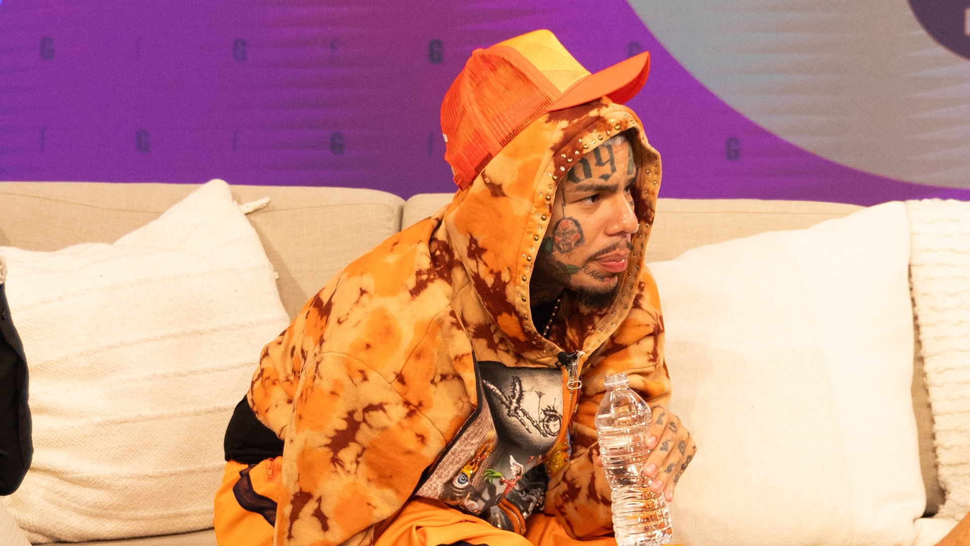 6ix9ine wearing an orange tie-dye hoodie and cap, sitting on a couch holding a water bottle.