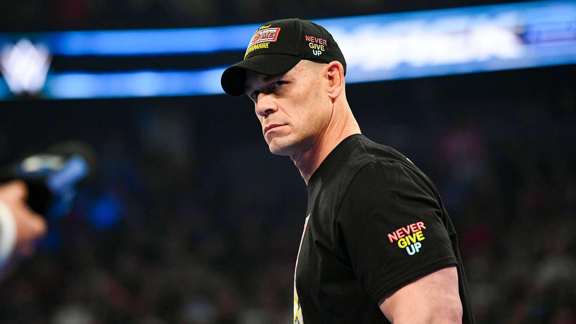 John Cena in a wrestling ring, wearing a "Never Give Up" cap and t-shirt, looking determined.