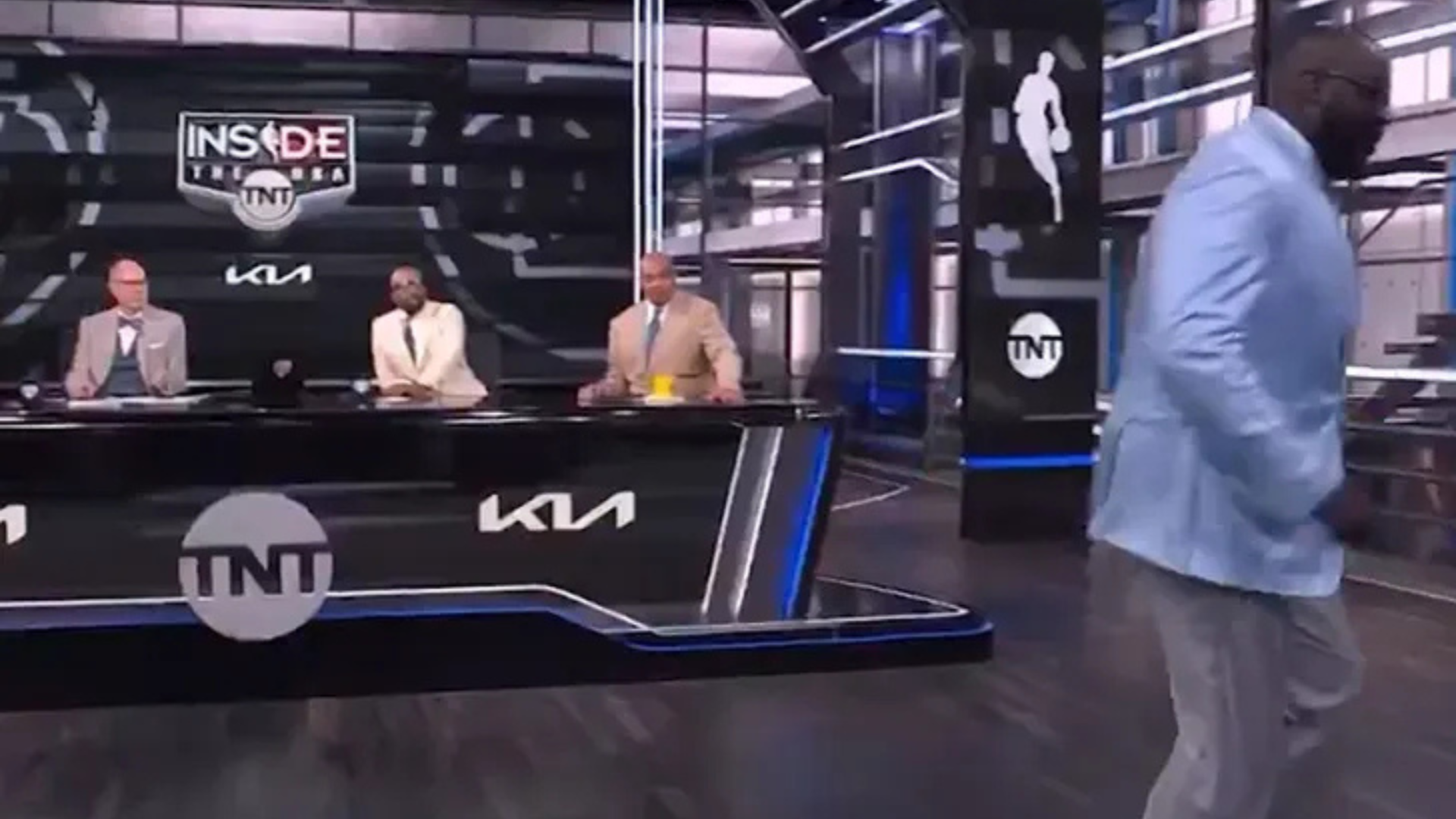 A TV studio with three men seated at a desk and one man in a blue blazer walking away. The set has "Inside the NBA" branding.