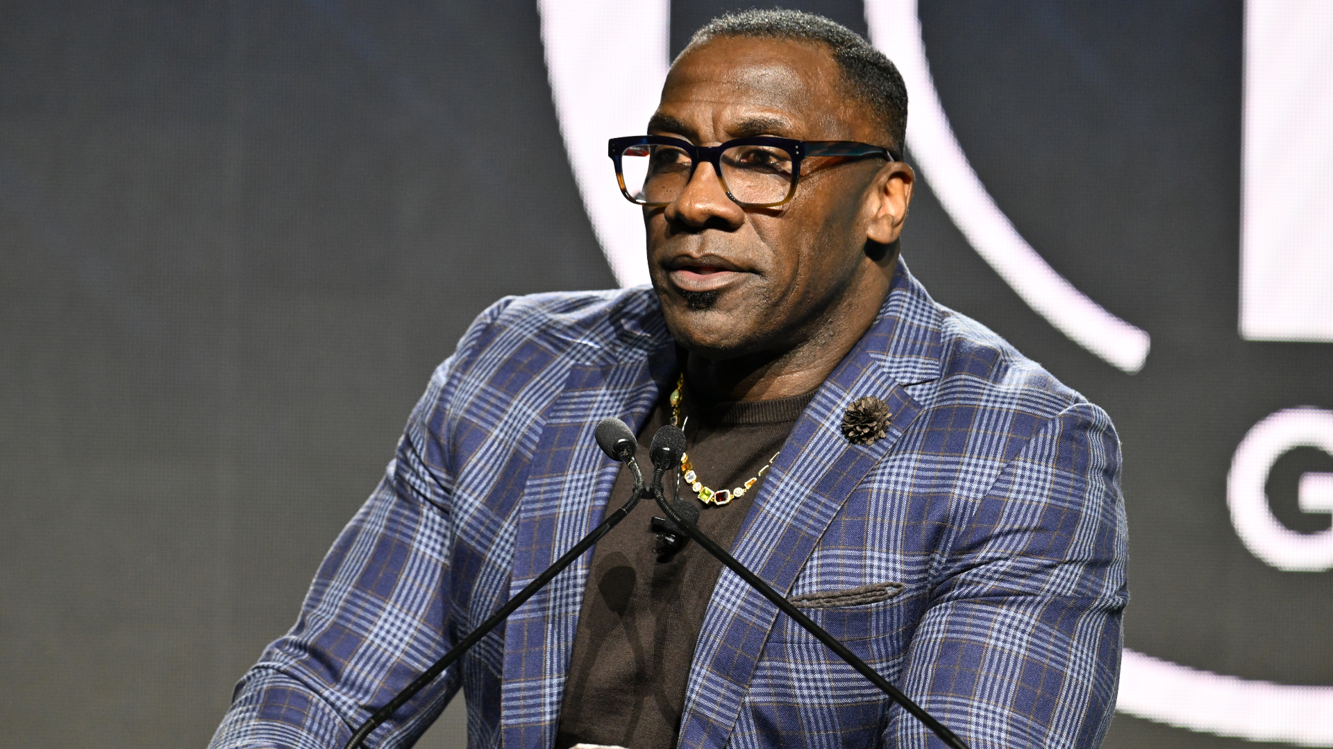 A man in glasses and a blue plaid jacket speaks at a podium.