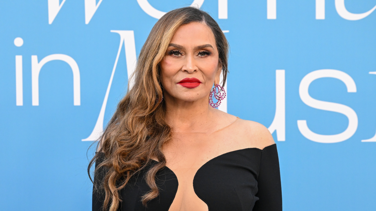 Tina Knowles with long wavy hair and red lipstick stands in front of a blue background with text. She's wearing a black dress.