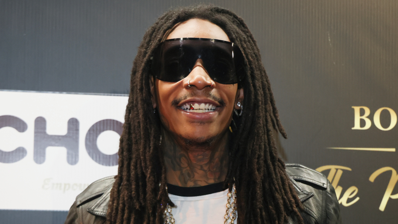 Rapper Wiz Khalifa wearing sunglasses and a leather jacket, smiling at an event.