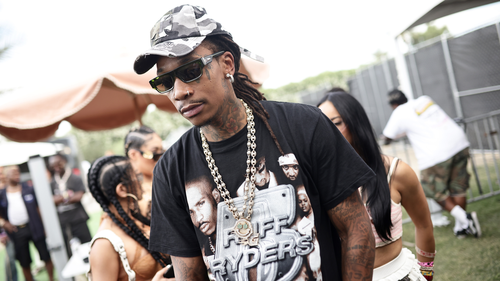 Wiz Khalifa wearing sunglasses, a camo cap, and a Ruff Ryders T-shirt, with chains around his neck, stands outdoors at an event.
