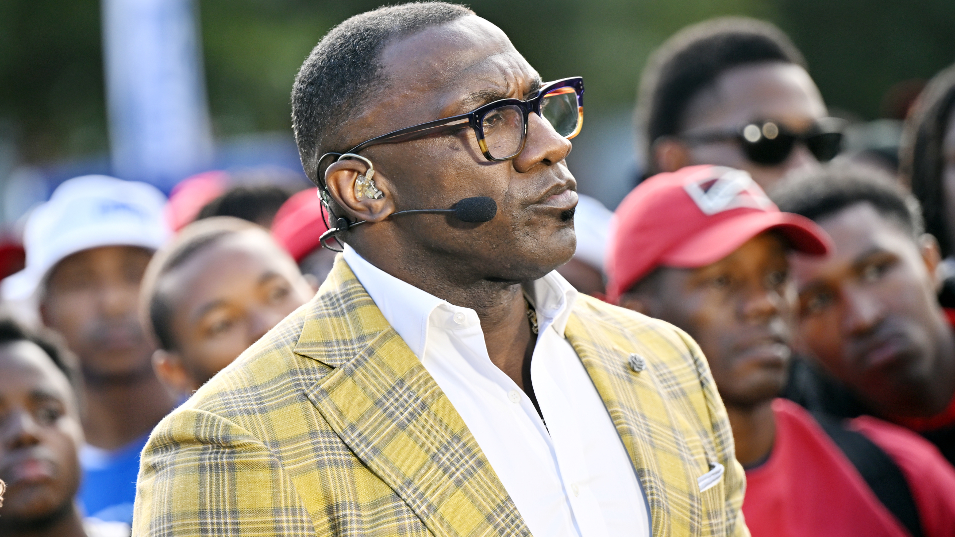 A man in a yellow plaid jacket and glasses, wearing a headset, stands among a crowd.