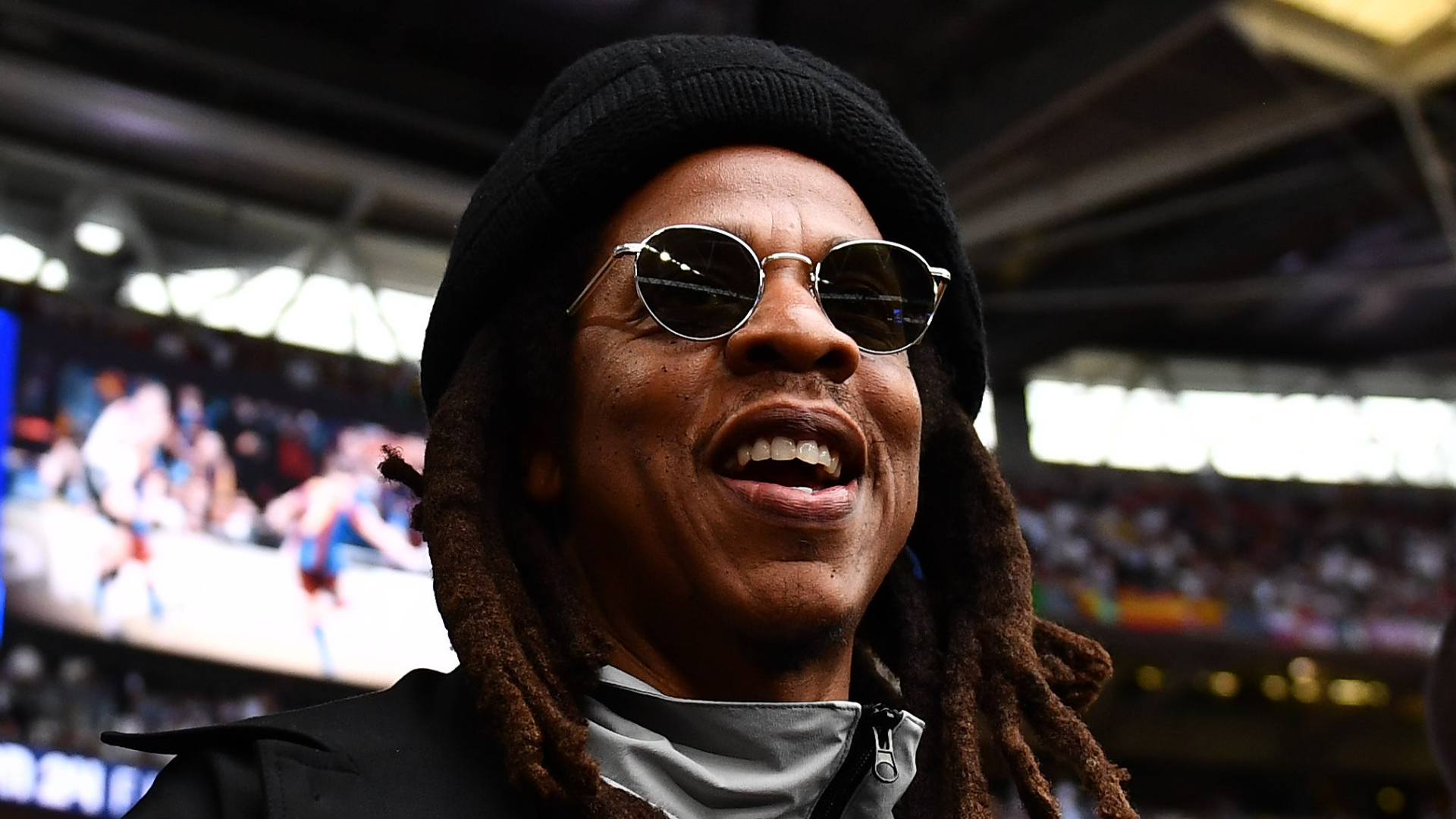 A person with long hair and sunglasses, wearing a black hat and coat, smiling in a stadium setting.