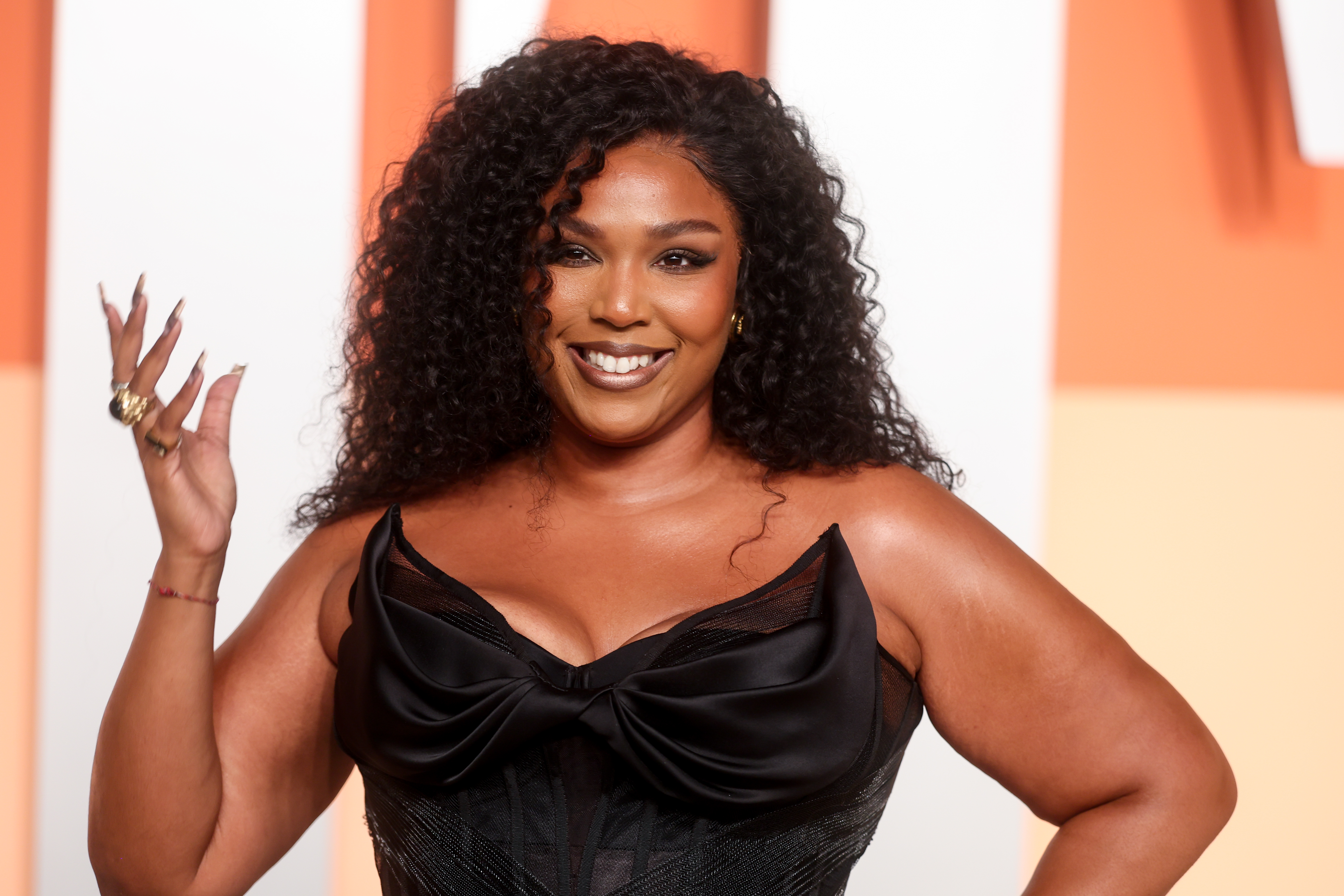 Lizzo poses in a black dress with a bow detail, smiling with her hand raised, against an orange and white background.