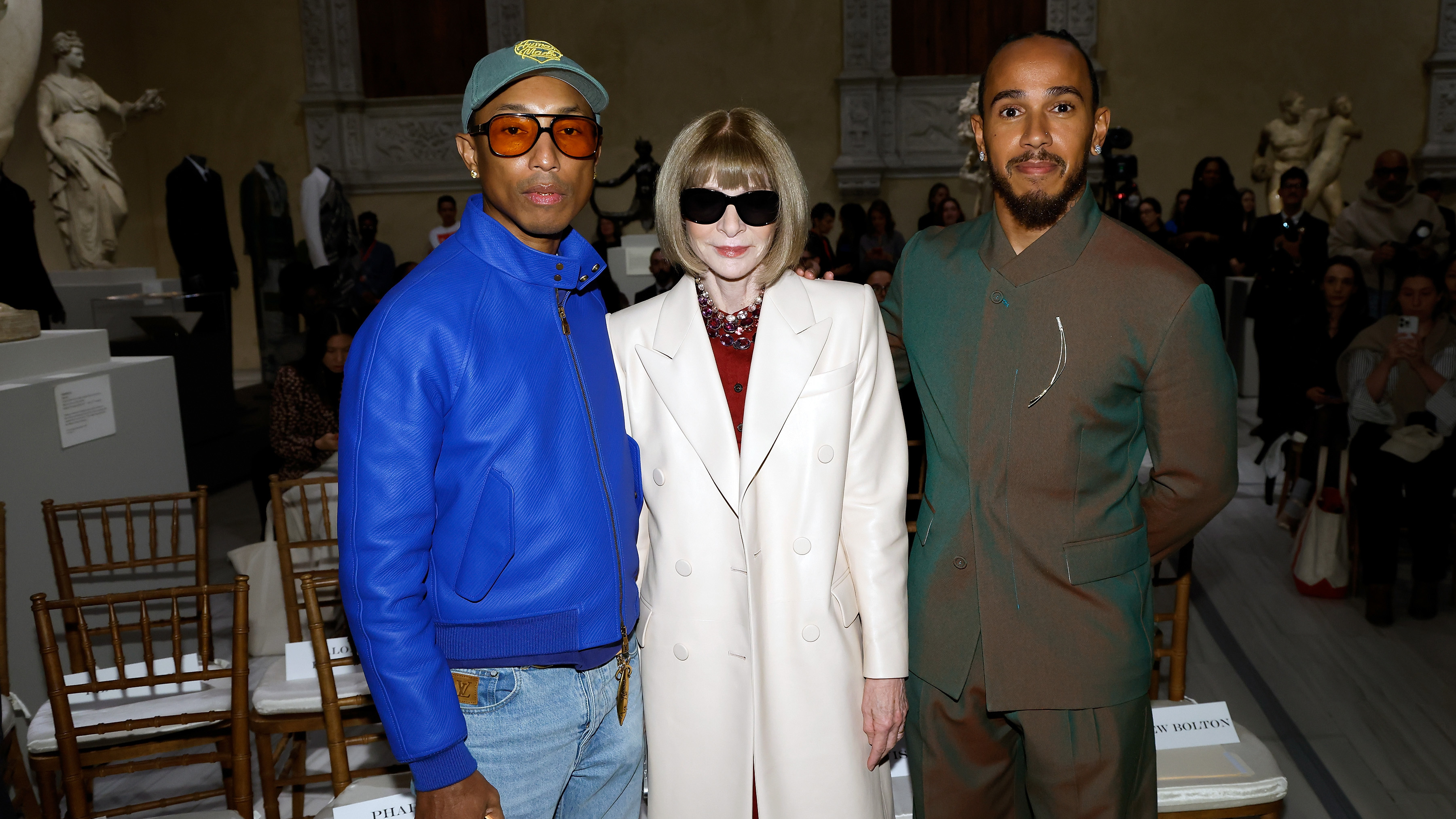 Pharrell Williams in a blue jacket, Anna Wintour in a white coat and sunglasses, and Lewis Hamilton in a brown suit at a fashion event for the 2025 Met Gala.