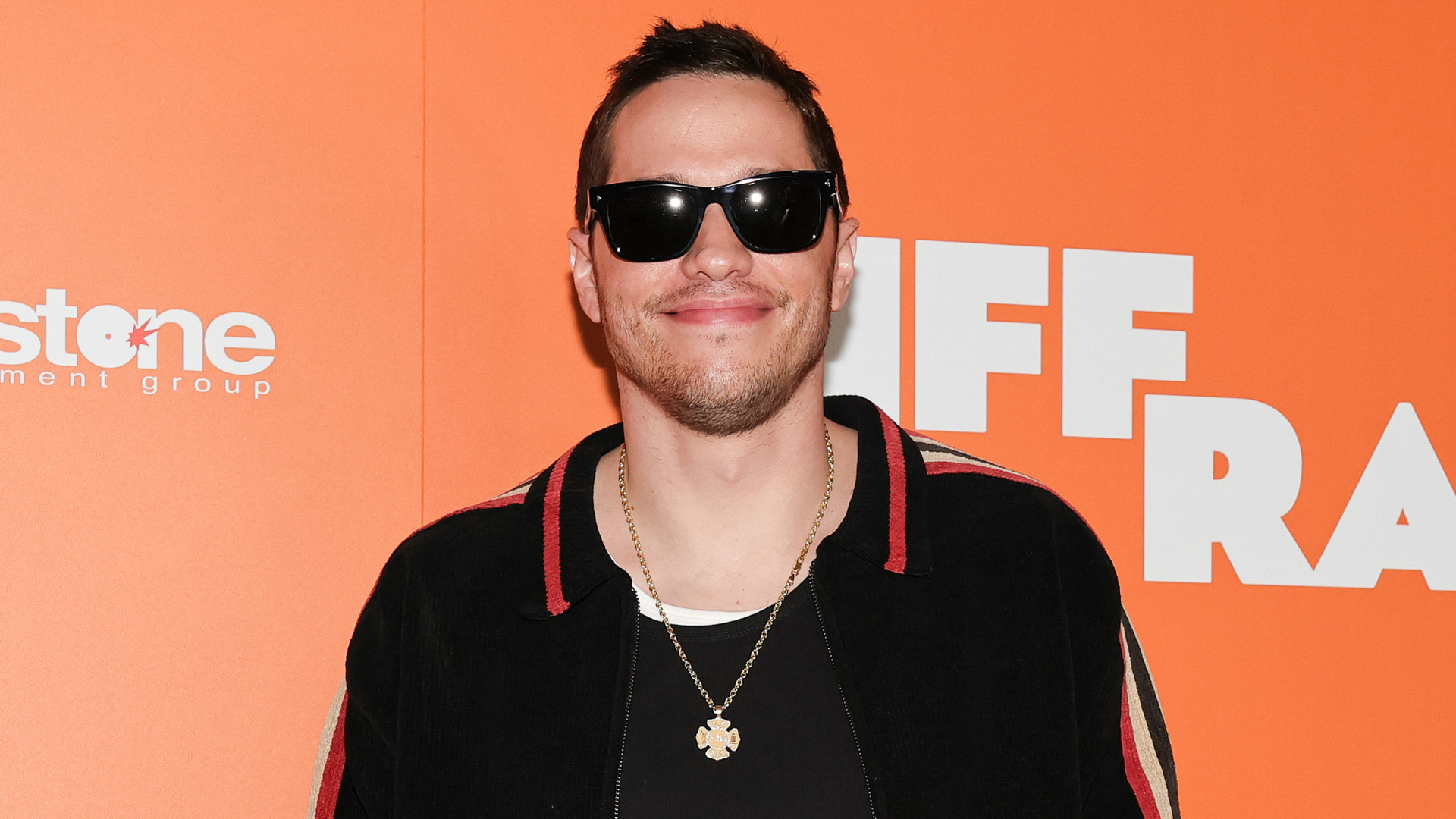 Pete Davidson wearing sunglasses and a black jacket, smiling against an orange background.