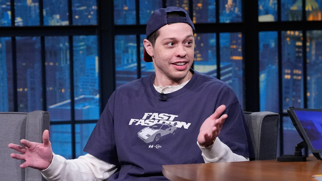 Pete Davidson wearing a t-shirt and cap, gesturing while seated on a talk show set.