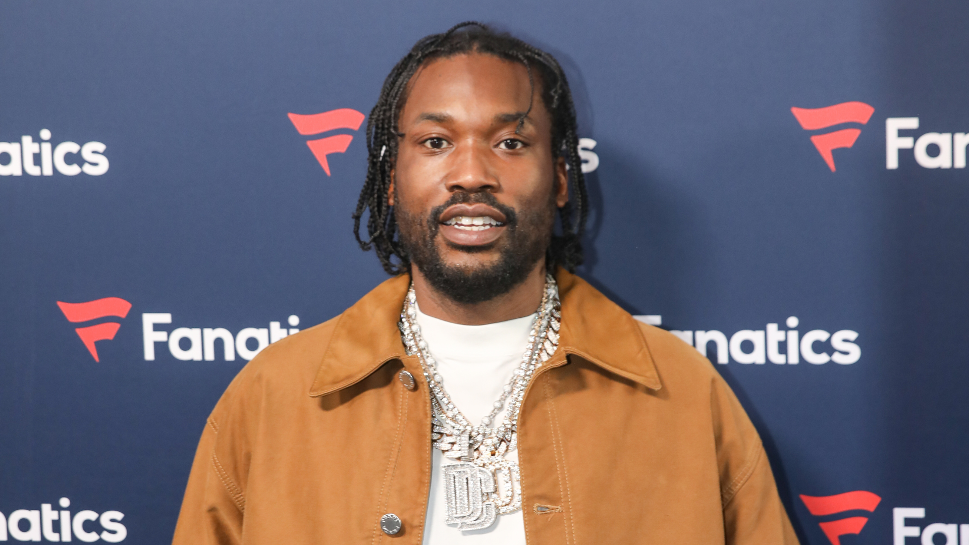 A man with braided hair and a brown jacket stands in front of a Fanatics logo backdrop, wearing a large chain necklace.