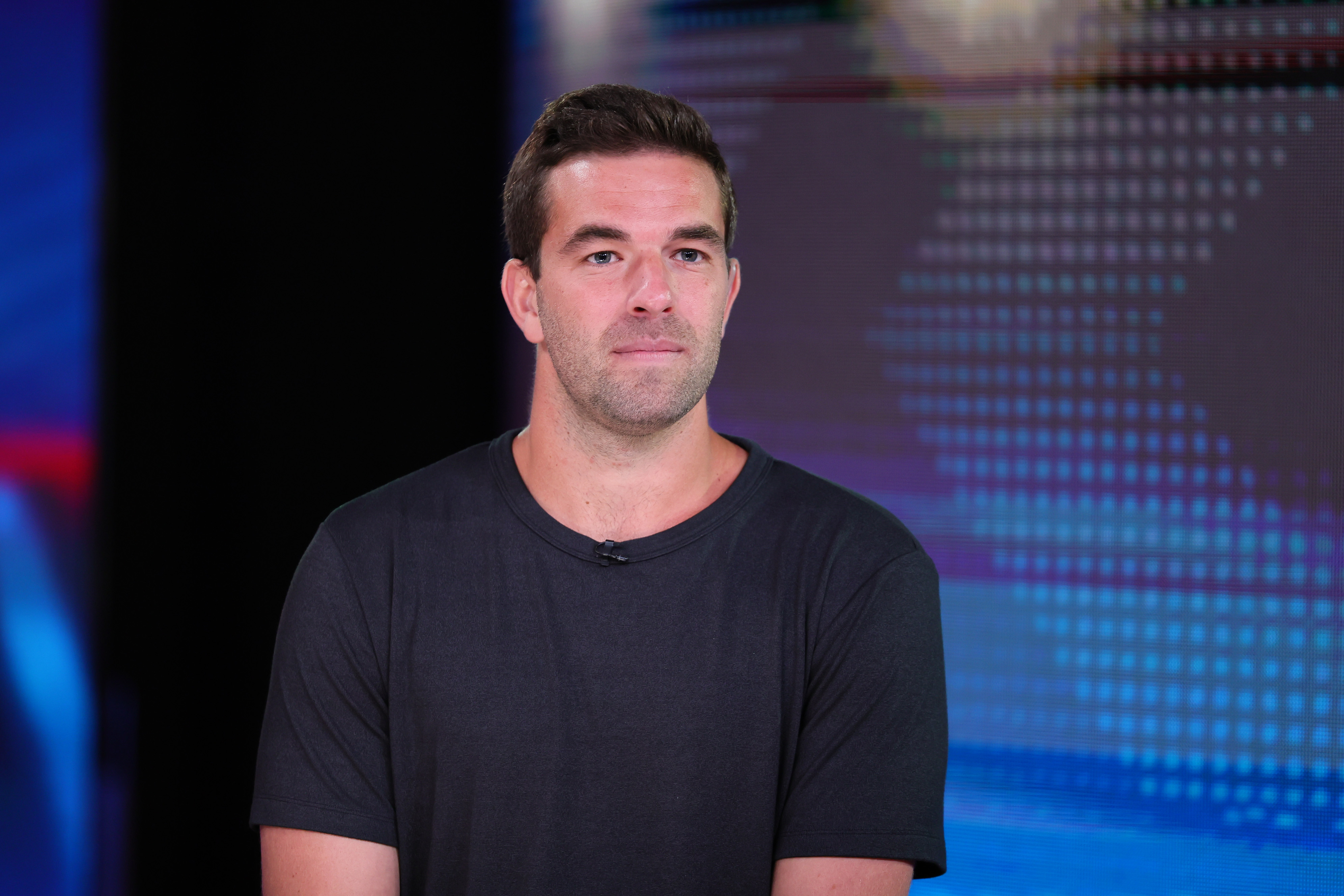 Fyre Festival founder Billy McFarland in a black t-shirt is standing in front of a digital screen with a dotted pattern.