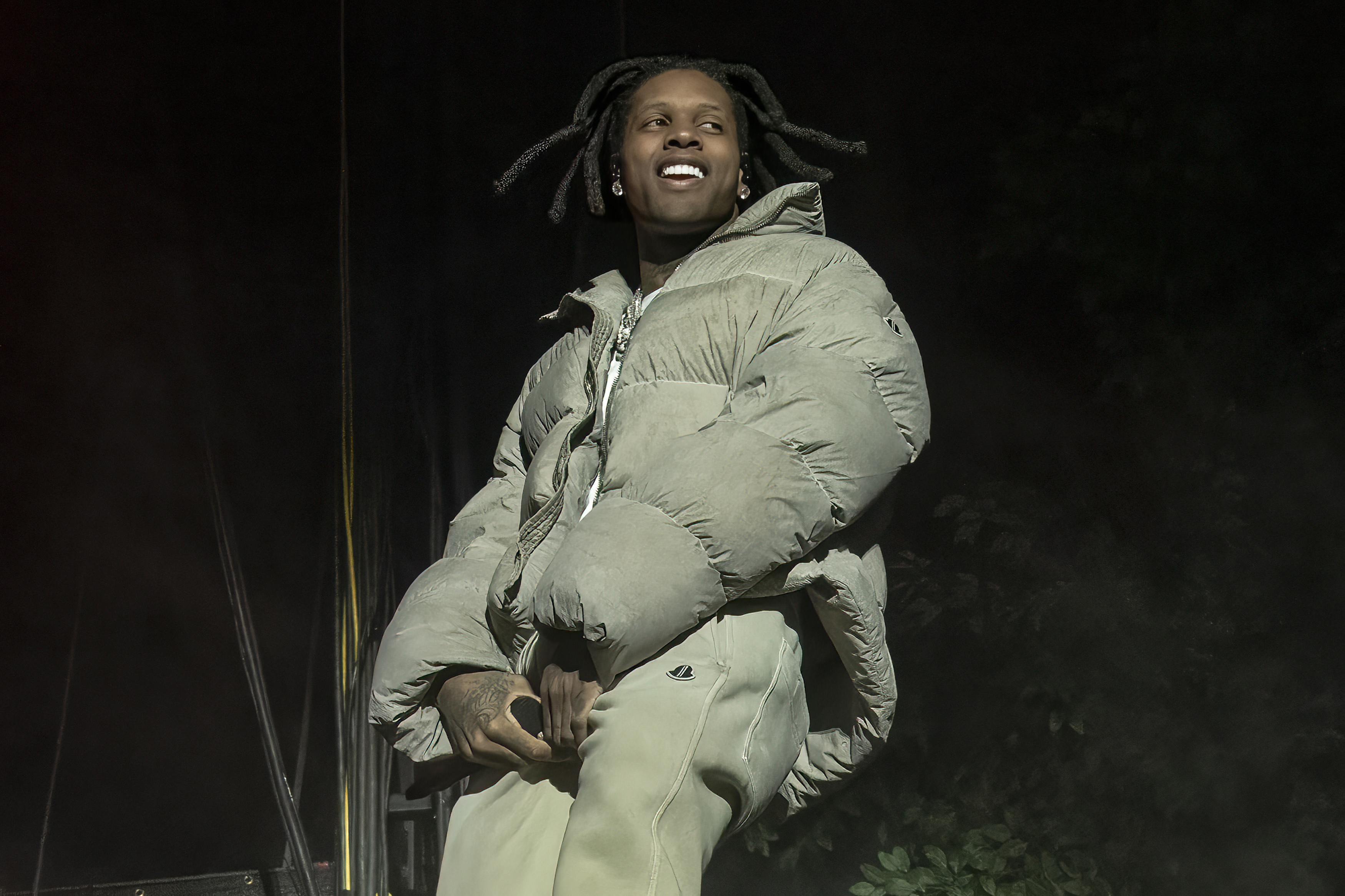 Lil Durk wearing a puffy jacket and pants, smiling on stage with a microphone in hand, against a dark background.