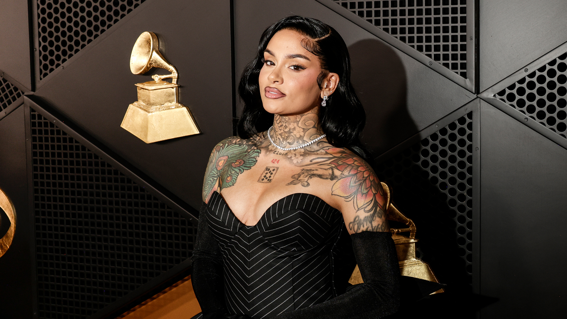 Kehlani at an event, wearing a pinstripe dress. She has visible tattoos and is posing in front of Grammy-themed decor.