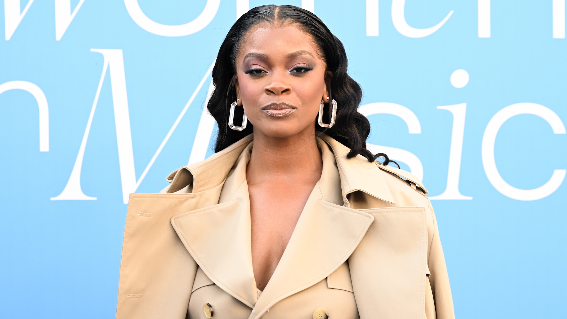 Ari Lennox poses in a beige trench coat against a blue background with text. She wears large hoop earrings and has wavy hair.