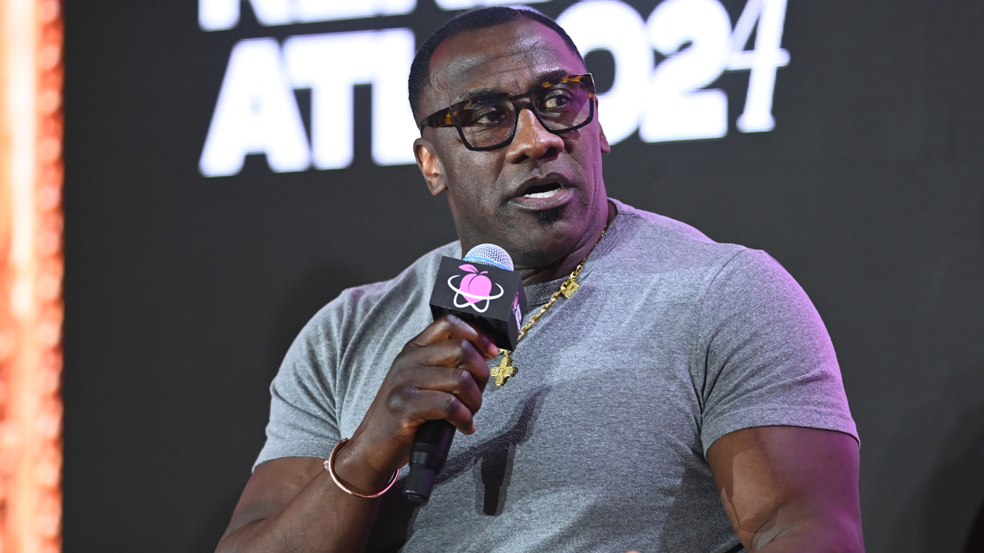 Shannon Sharpe in glasses and a gray shirt speaks into a microphone on stage, with "2024" visible in the background.