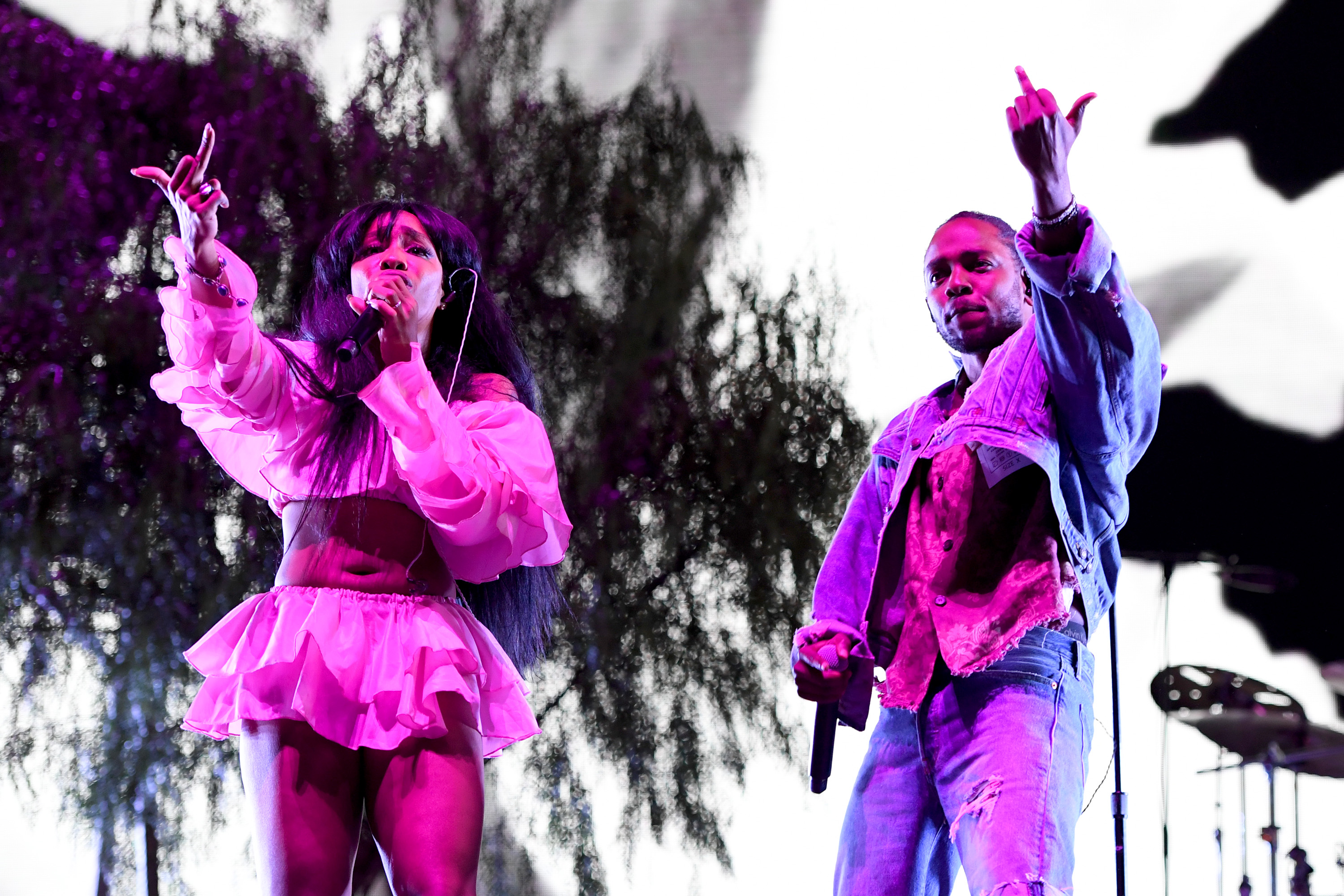 SZA and Kendrick Lamar performing on stage, both wearing casual outfits and holding microphones, with expressive gestures.