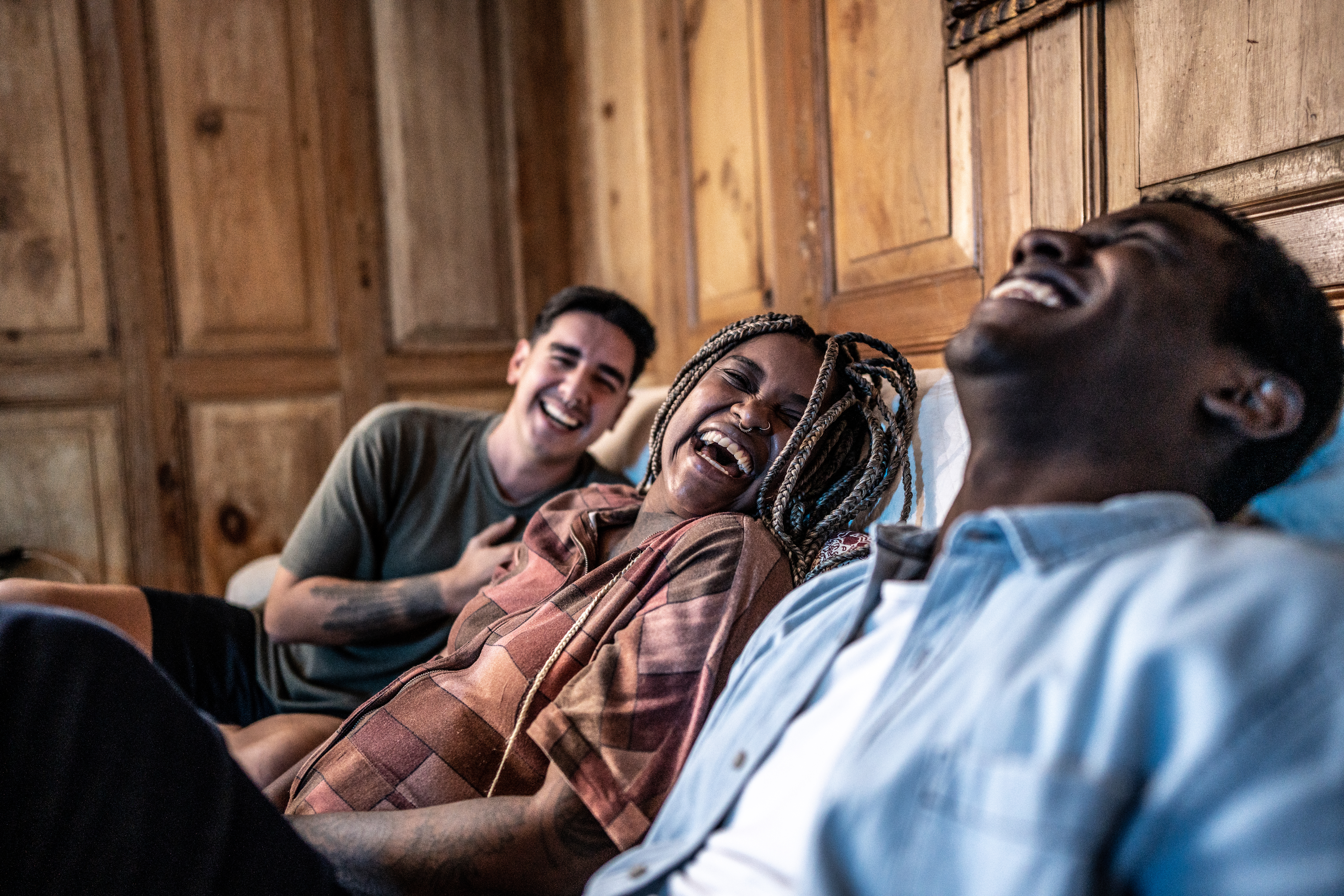 Three people sitting on a couch, laughing together in a warmly lit room with wooden paneling.