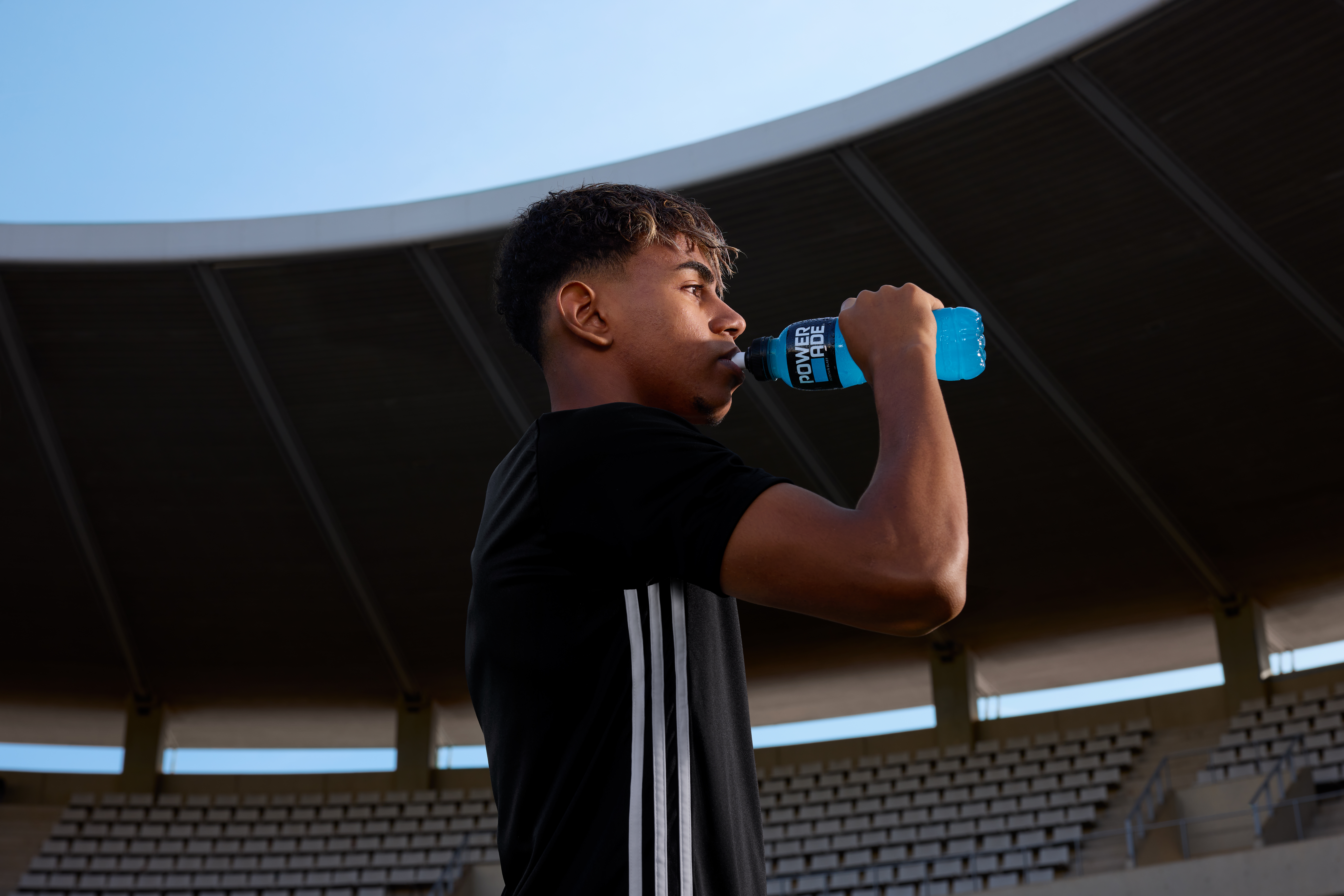 A person in a black shirt drinks from a blue bottle labeled "Prime" in a stadium with empty seats.