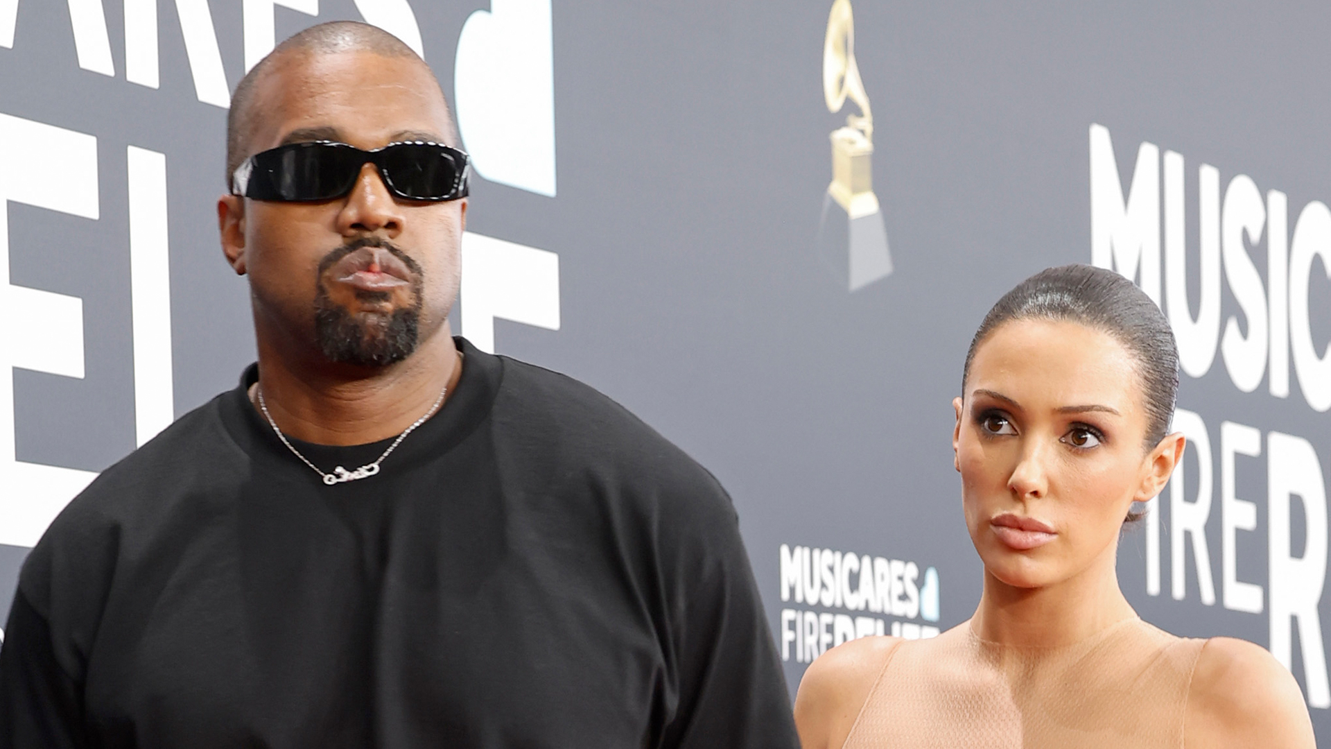 Kanye West wearing sunglasses and a black shirt stands beside Bianca Censori in a beige outfit at an event with a black backdrop.