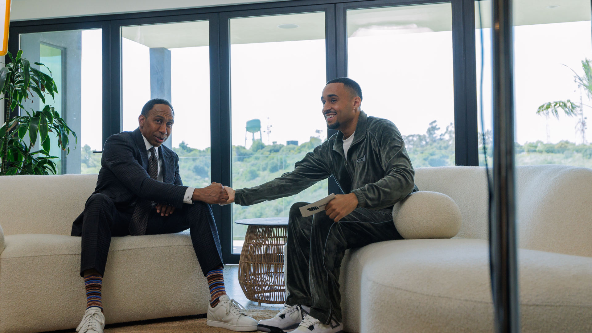 Stephen A. Smith and Speedy Morman shaking hands on a sofa, one in a suit and the other in a casual outfit, with a scenic view outside the window.