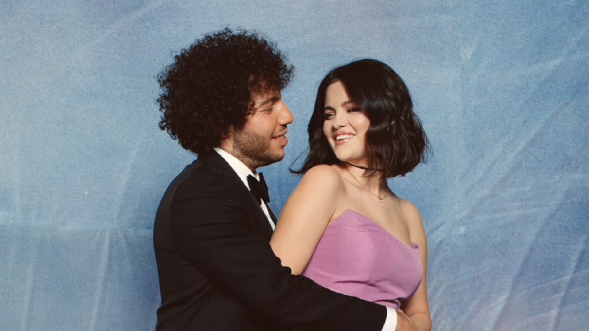 Benny Blanco in a suit embraces Selena Gomez in a purple dress against a blue background. They are smiling at each other.
