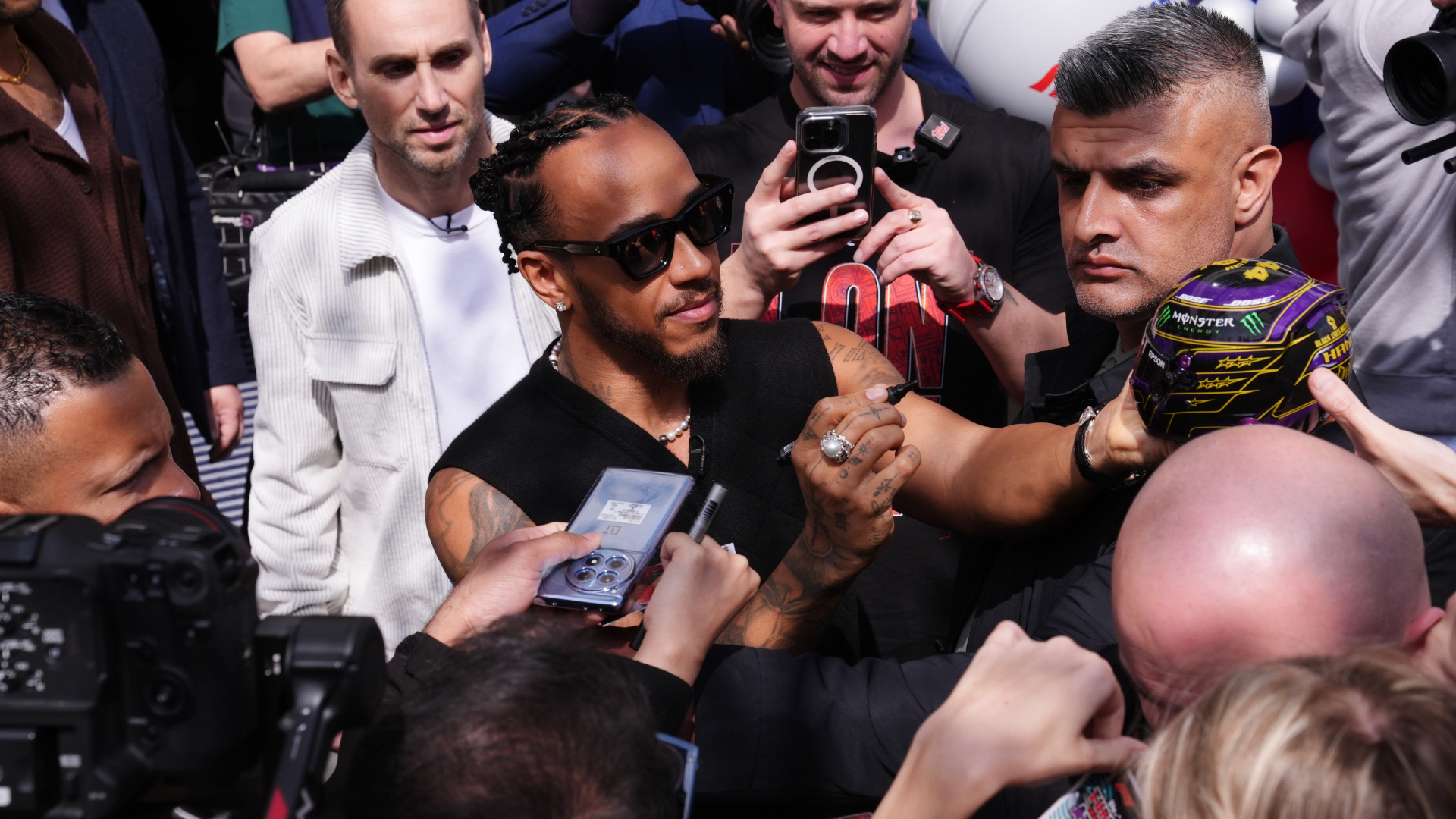 Lewis Hamilton, wearing sunglasses and a sleeveless top, interacts with fans and signs autographs in a crowded setting.