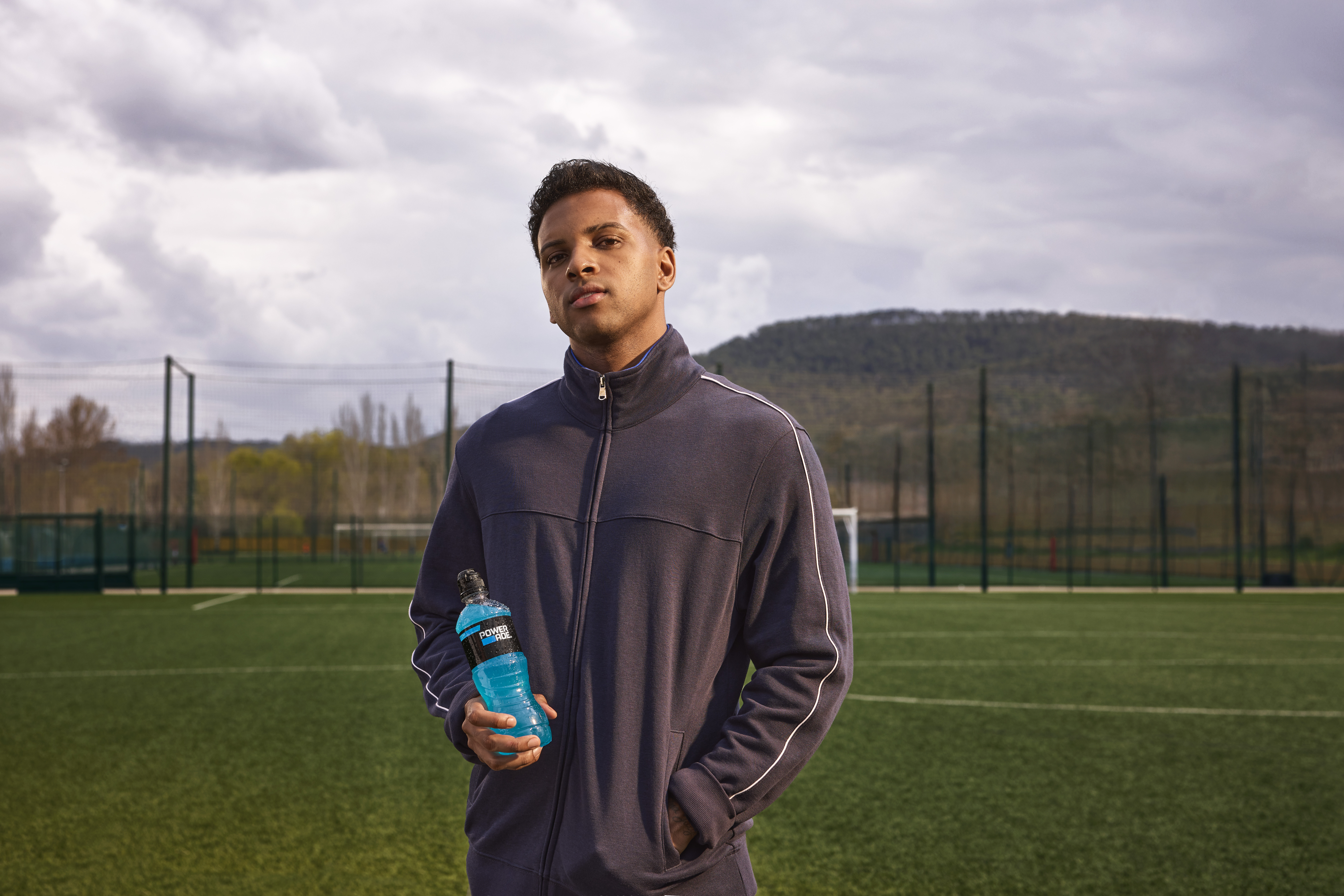 Rodrygo Goes in a track jacket stands on a soccer field, holding a blue sports drink bottle. The background shows a fenced field and hills.