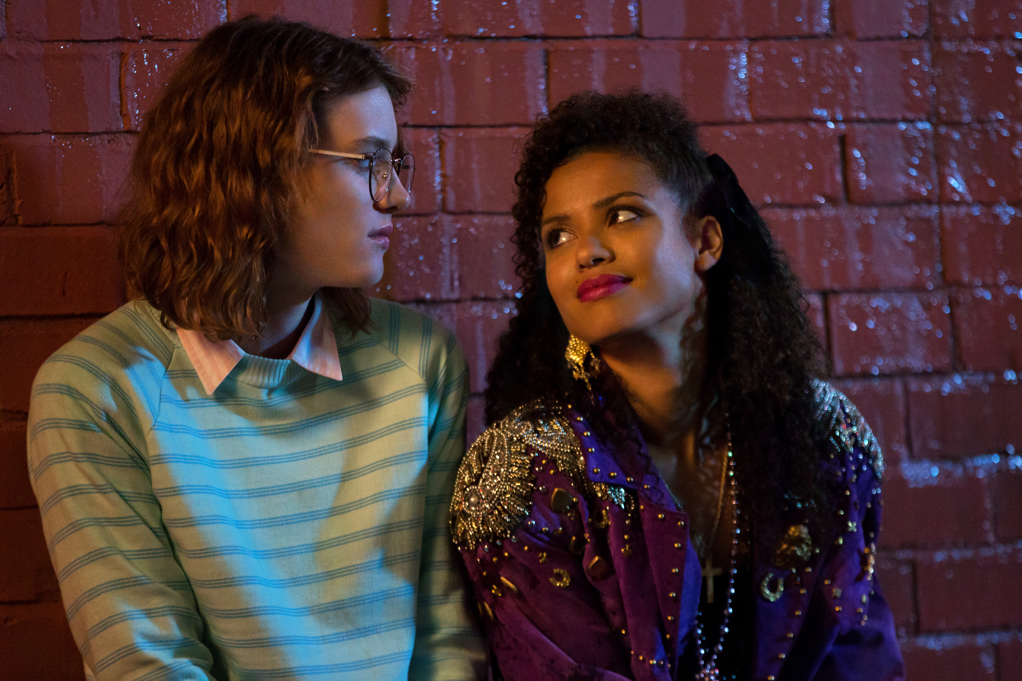 Two women sit against a brick wall. One wears glasses and a striped sweater, the other a purple embellished jacket, looking at each other.