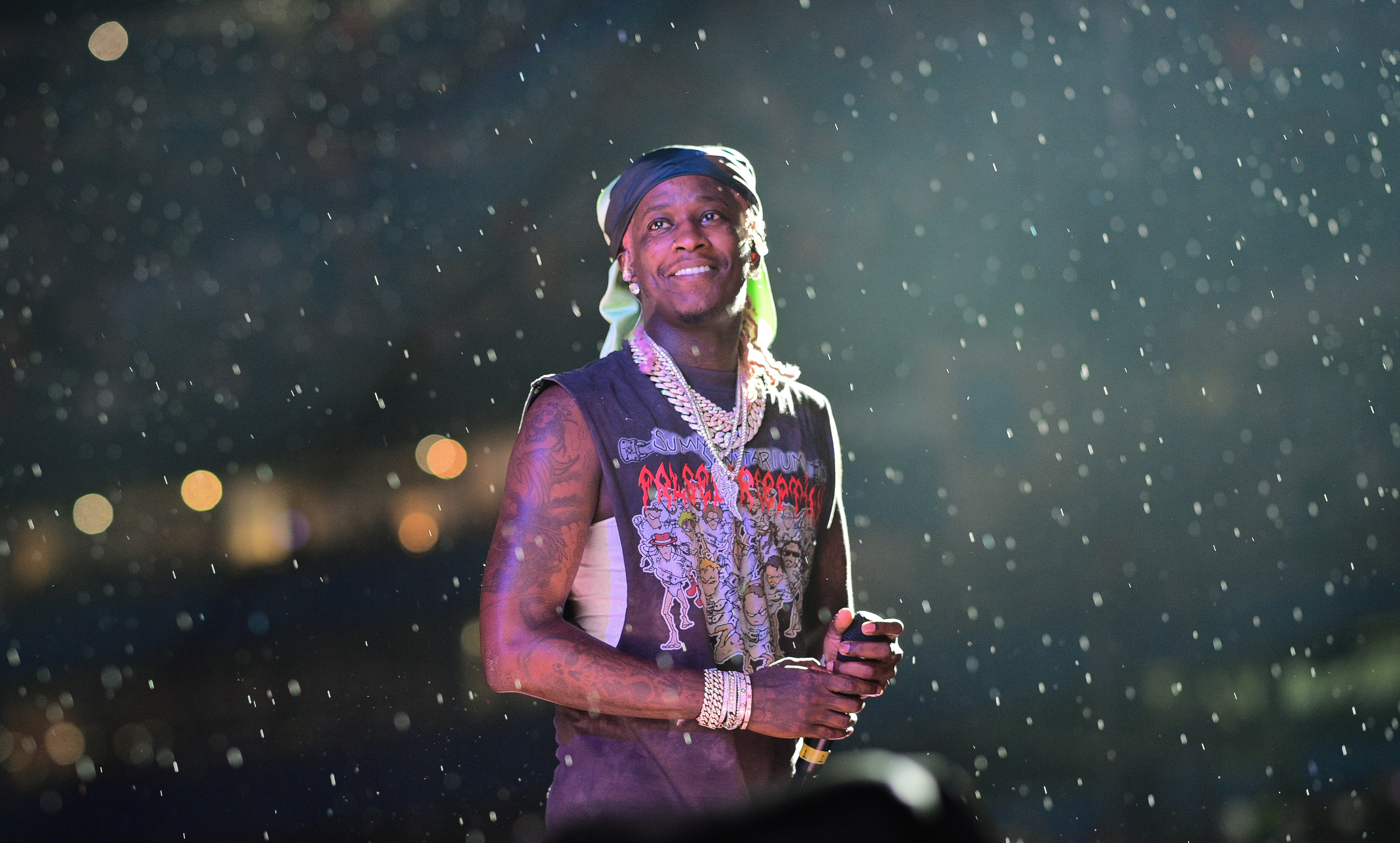 Young Thug on stage in the rain, wearing a graphic tank top, bandana, and jewelry, holding a microphone, with blurred lights in the background.
