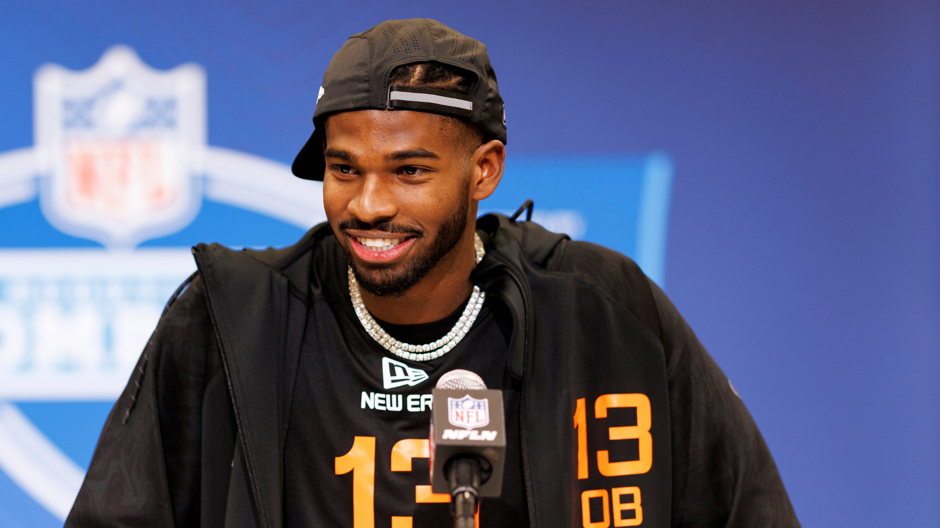 A man wearing a black cap and jacket with the number 13, smiling at a press conference with an NFL backdrop.