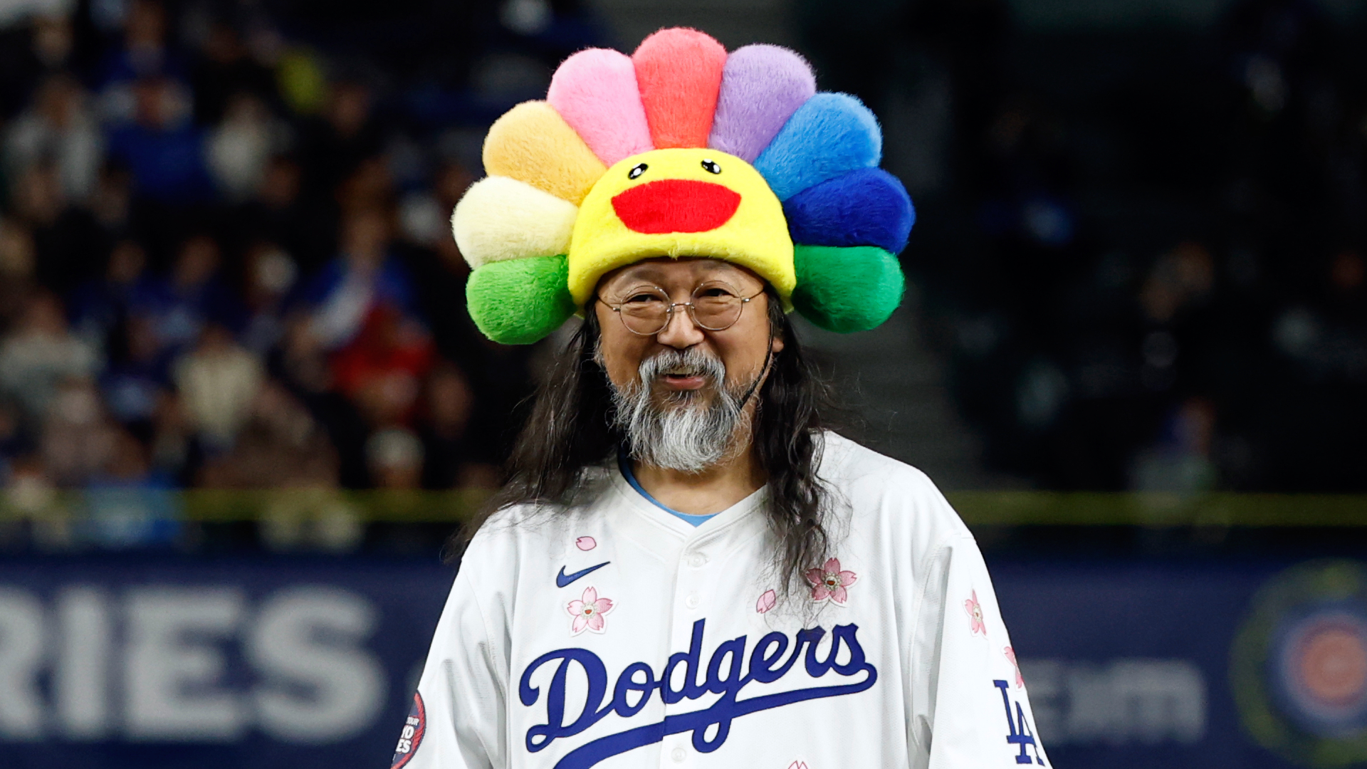 Artist Takashi Murakami wearing a colorful flower hat and Dodgers jersey, smiling at a baseball game.