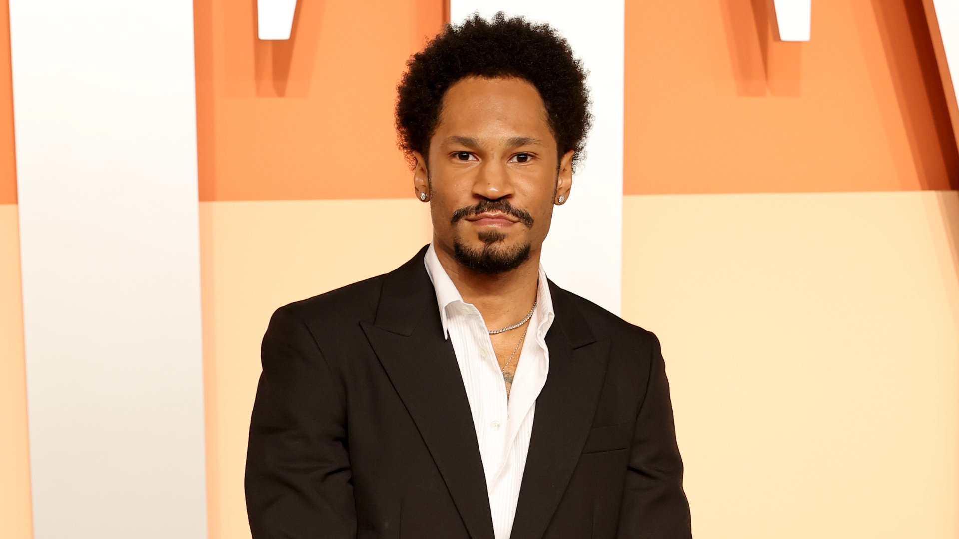 A man with short curly hair and a goatee, wearing a black suit and white shirt, stands against an orange and white background.