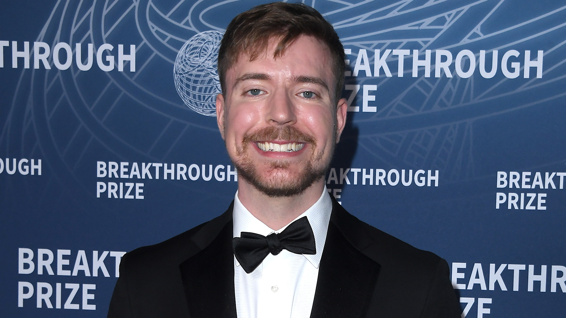 MrBeast smiles at the Breakthrough Prize event, with a blue background featuring the event's logo.