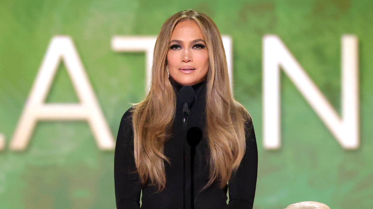 Jennifer Lopez speaking at an event, wearing a black outfit, with long wavy hair, against a green background.