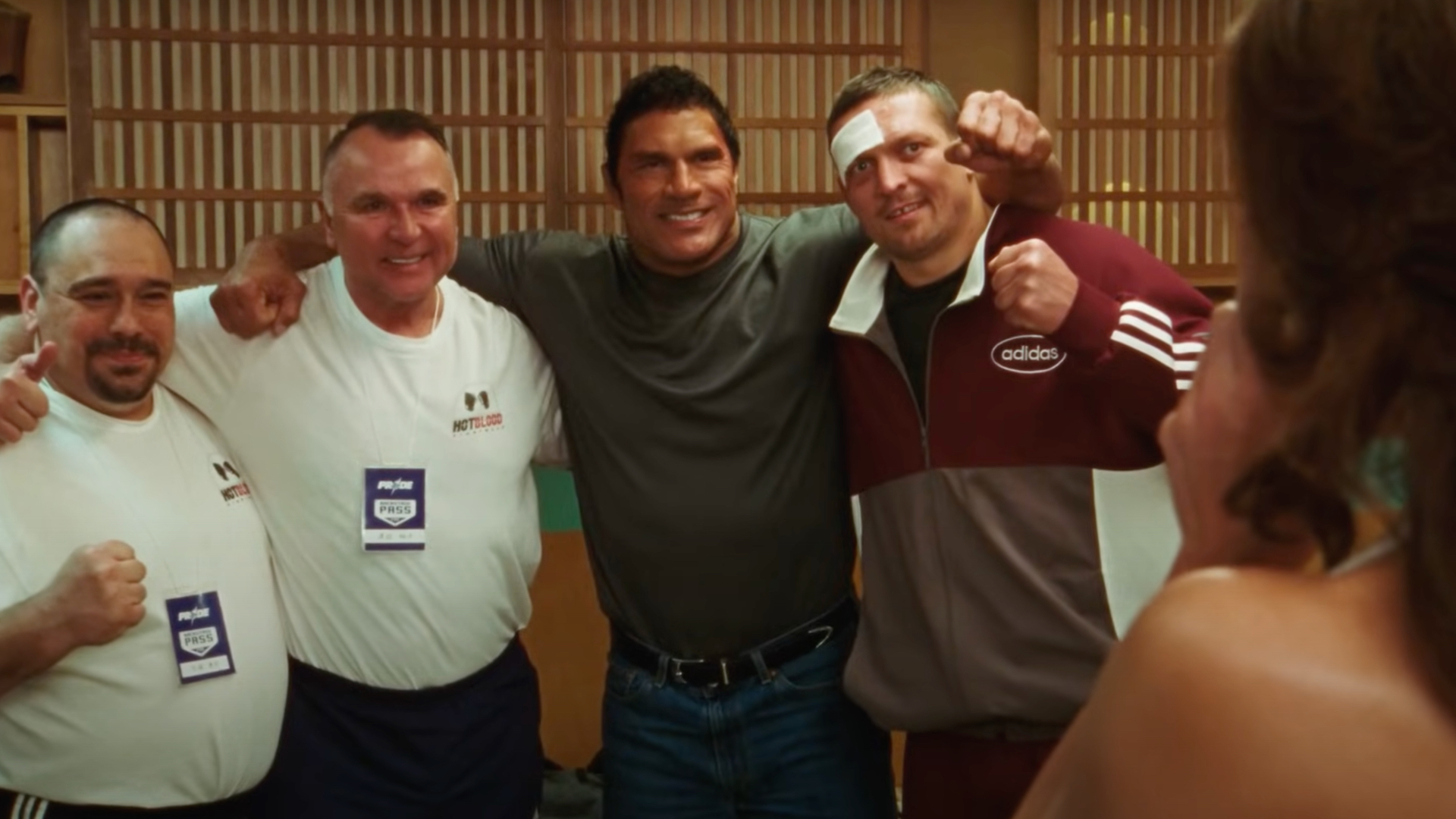 Four men posing together, one wearing an Adidas tracksuit and another in a grey shirt, all smiling and holding fists up.
