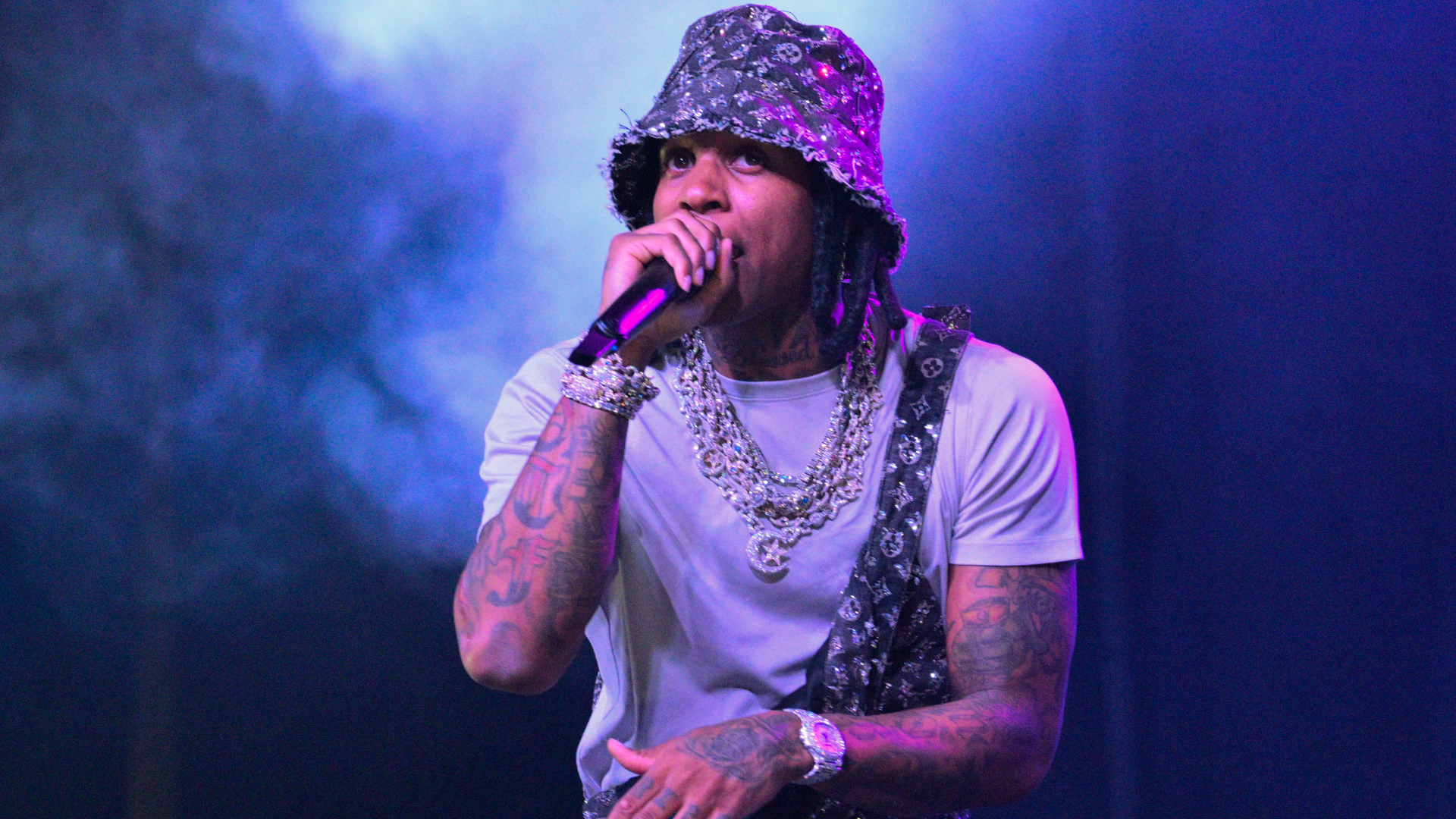 Lil Durk performing on stage, wearing a patterned bucket hat and chains, holding a microphone with a smoky purple background.