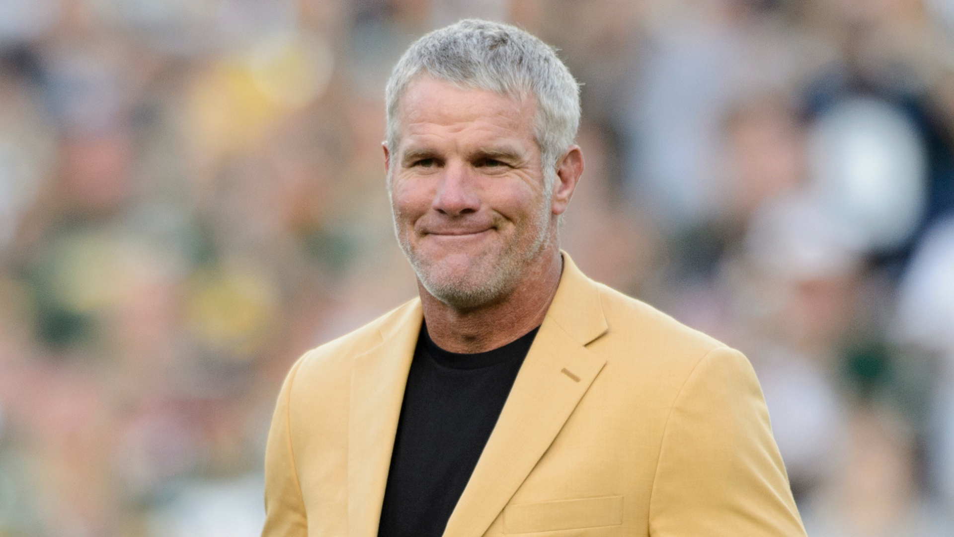 A man with short gray hair, wearing a yellow jacket and black shirt, standing outdoors with a blurred crowd in the background.