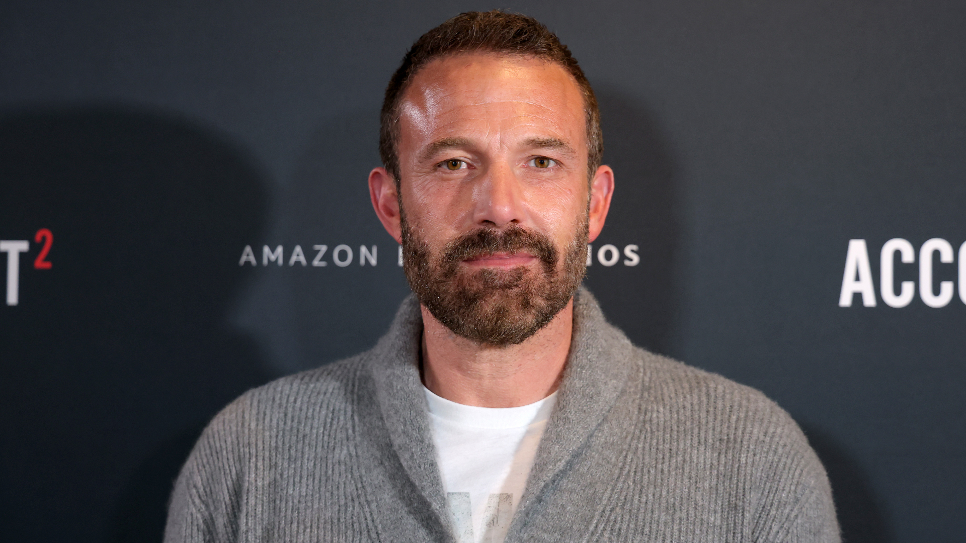 Ben Affleck at an event, wearing a gray cardigan and white shirt, standing against a backdrop with Amazon Studios branding.