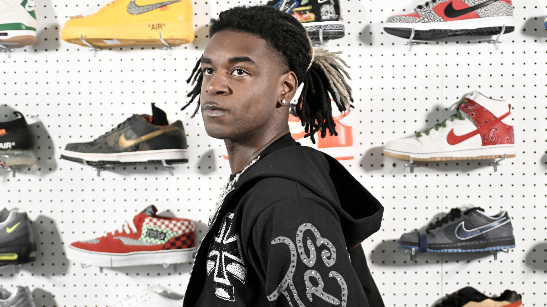 A person with braided hair stands in front of a wall displaying various colorful sneakers, wearing a black hoodie with designs.