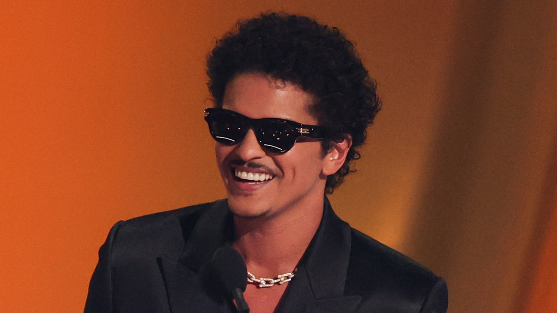 Bruno Mars, wearing sunglasses and a dark suit, smiles at an event.