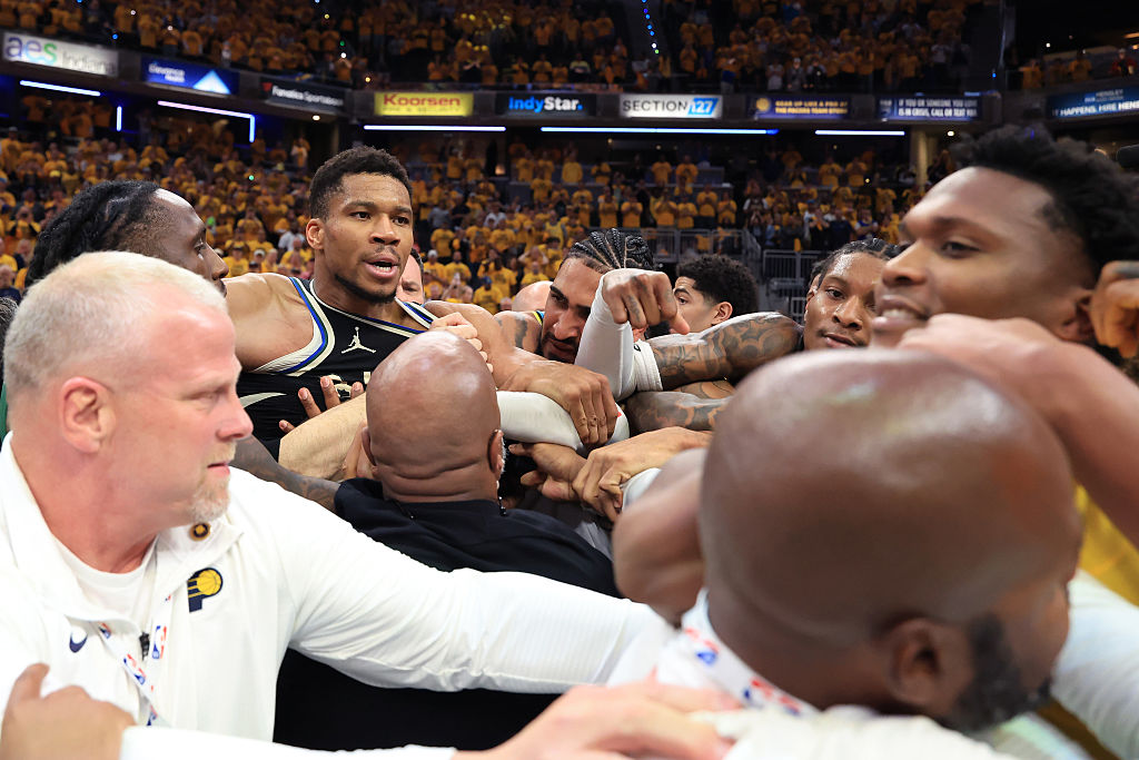 A group of basketball players and staff, including Giannis Antetokounmpo, in a huddle on a court surrounded by a cheering crowd.