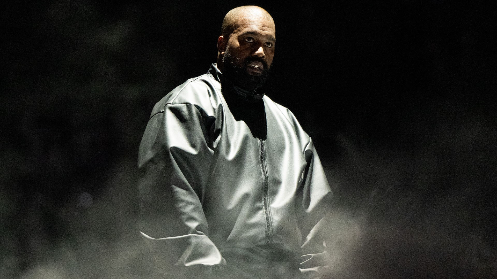 Kanye West in a dark, oversized jacket stands in a dimly lit setting, surrounded by mist.