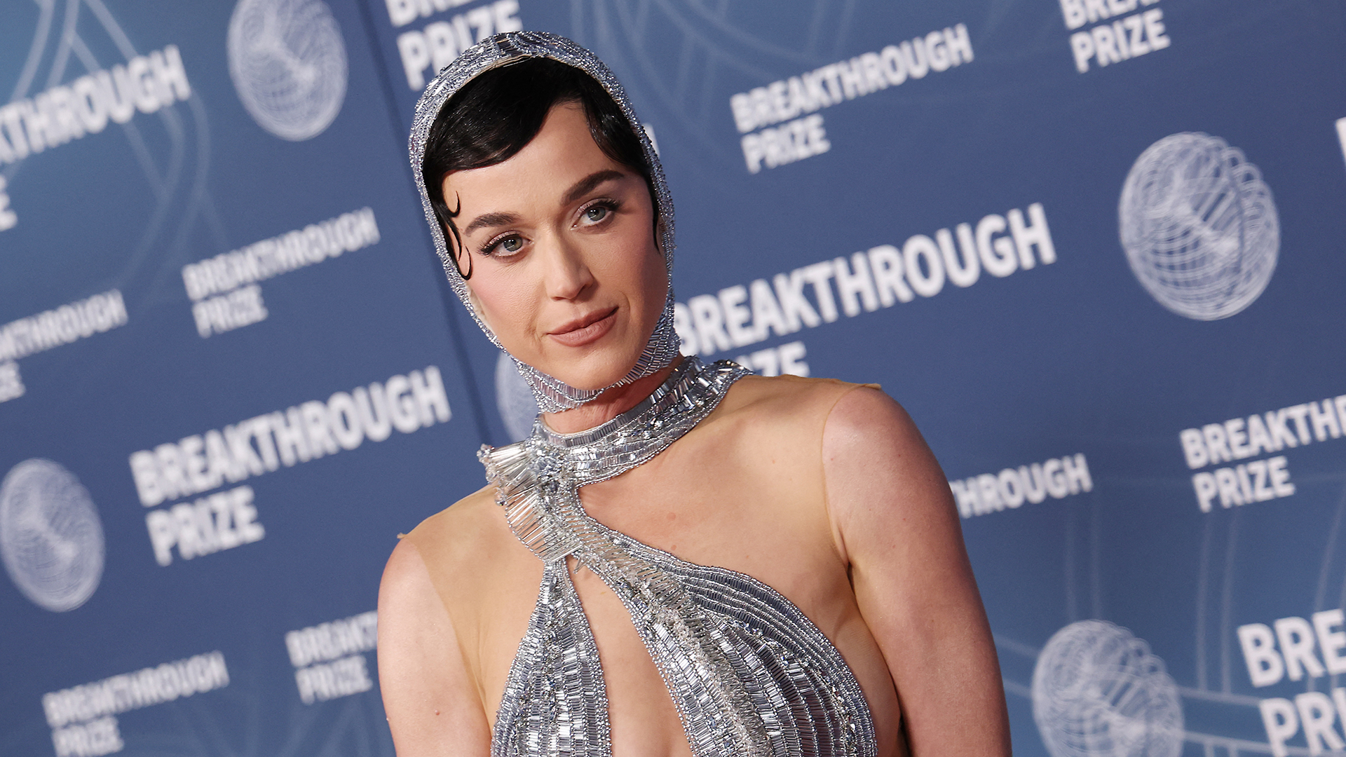 Katy Perry at the Breakthrough Prize event, wearing a sparkling silver gown with a matching headpiece, posing on the red carpet.